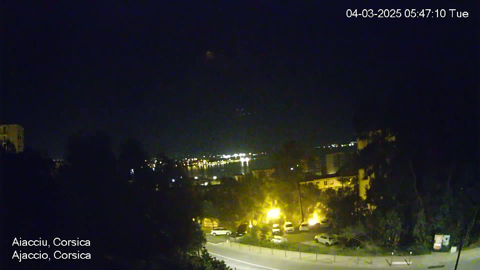 A nighttime view of a town by a body of water, with streetlights illuminating cars in a parking lot and a road below, under a clear, dark sky.