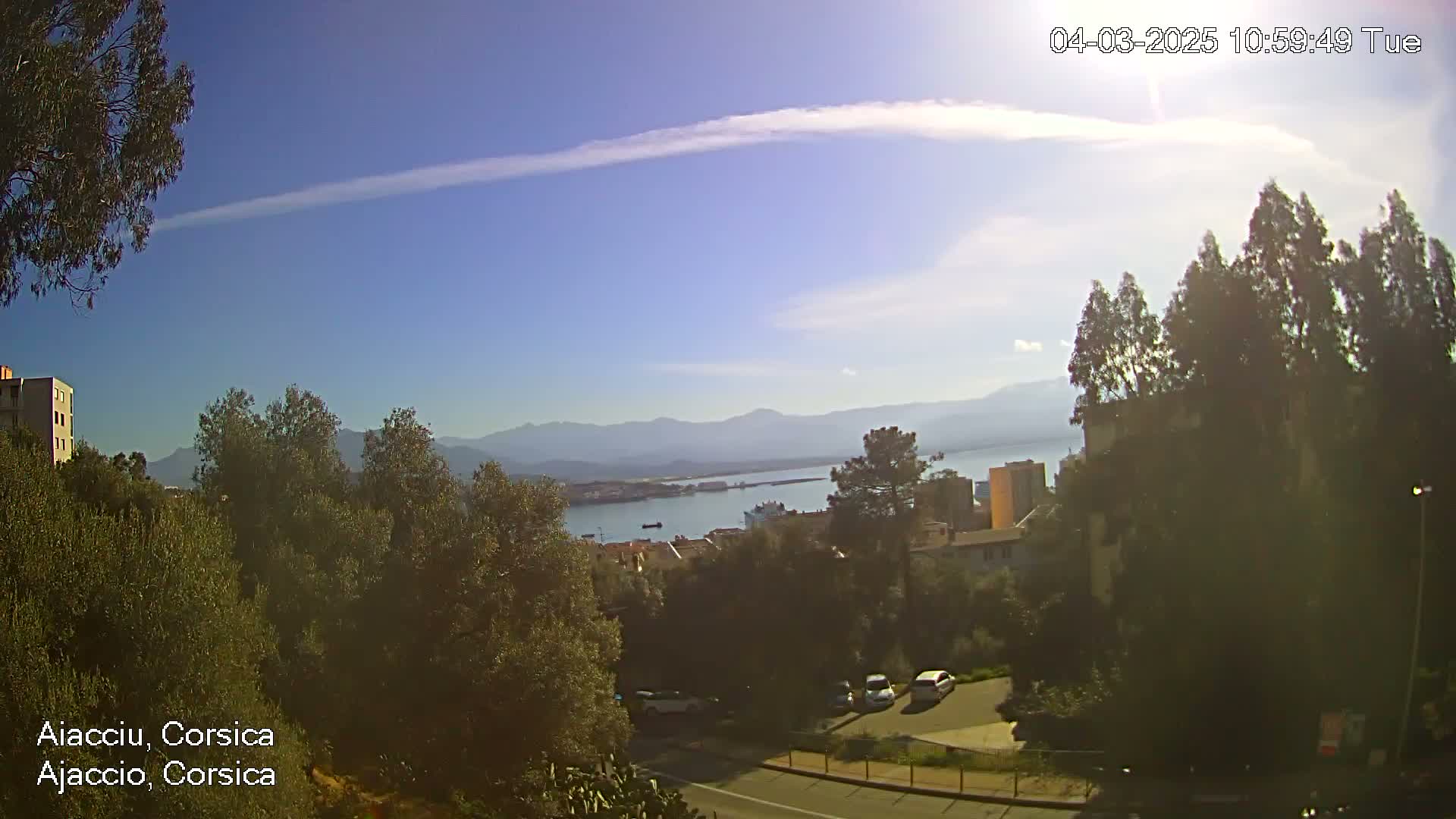 A partly sunny day reveals a coastal view of a town nestled among trees and hills, with a body of water and distant mountains in the background.