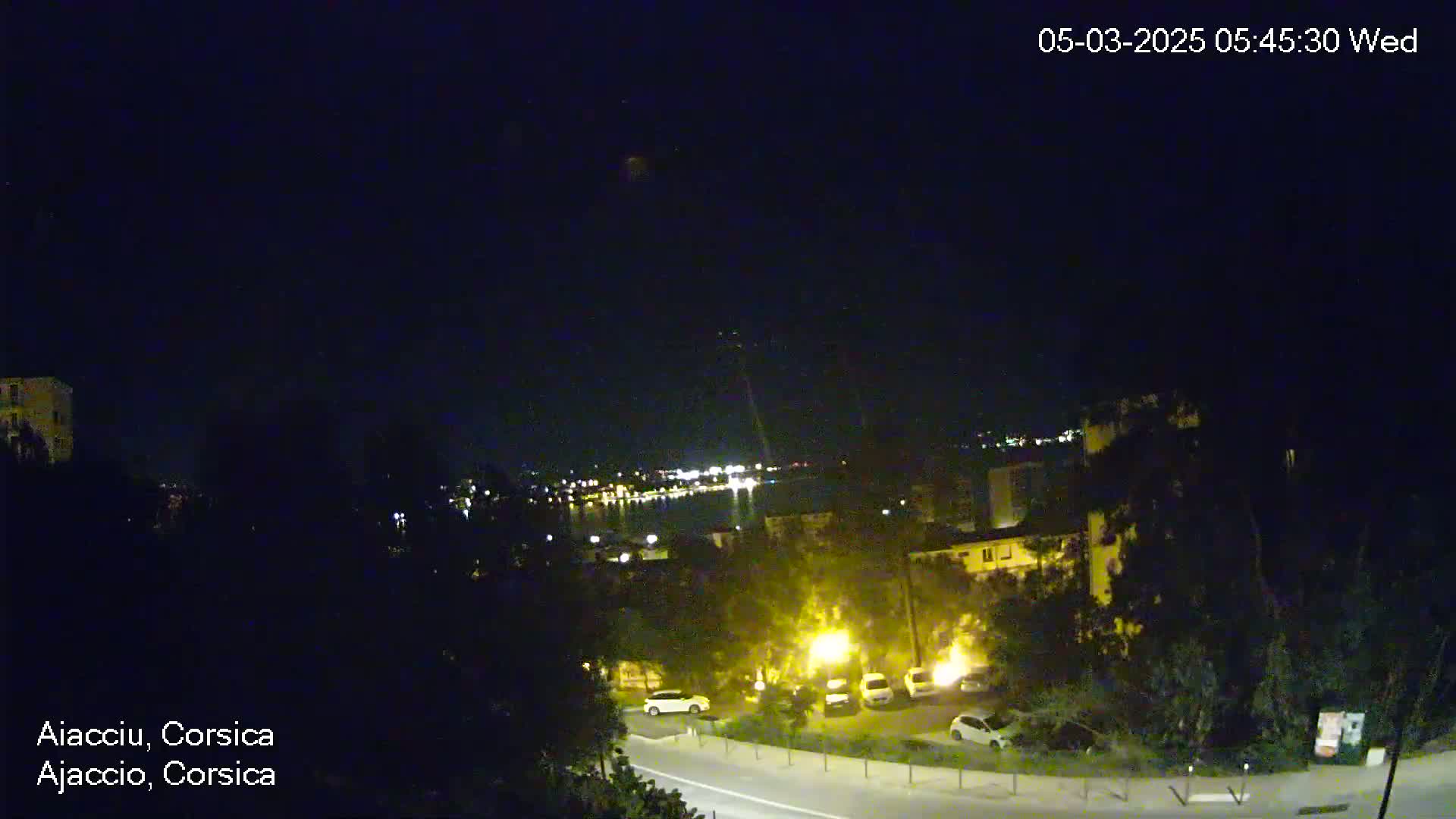 A nighttime, clear-sky view of a coastal town with dimly lit buildings, parked cars, and a road leading towards a body of water with lights in the distance.