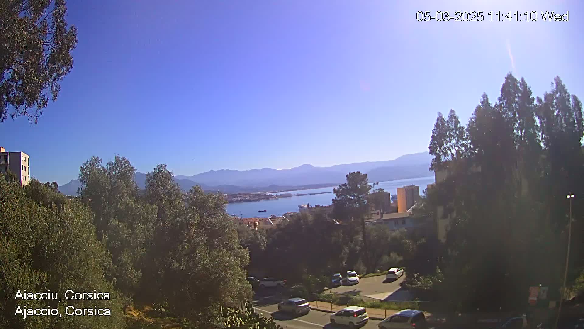 A clear, sunny day reveals a panoramic view of a coastal town nestled between mountains and a body of water, with cars visible on a road below.
