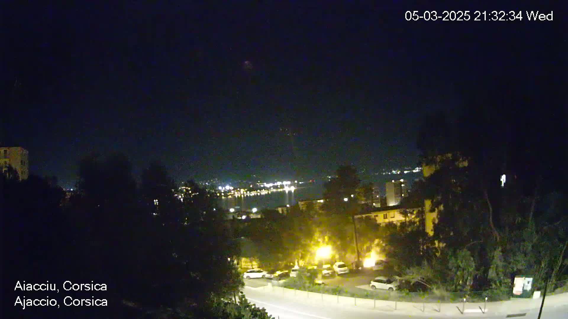 A nighttime view of a coastal city, illuminated with streetlights, overlooking a dark body of water with distant city lights across it under a clear, dark sky.