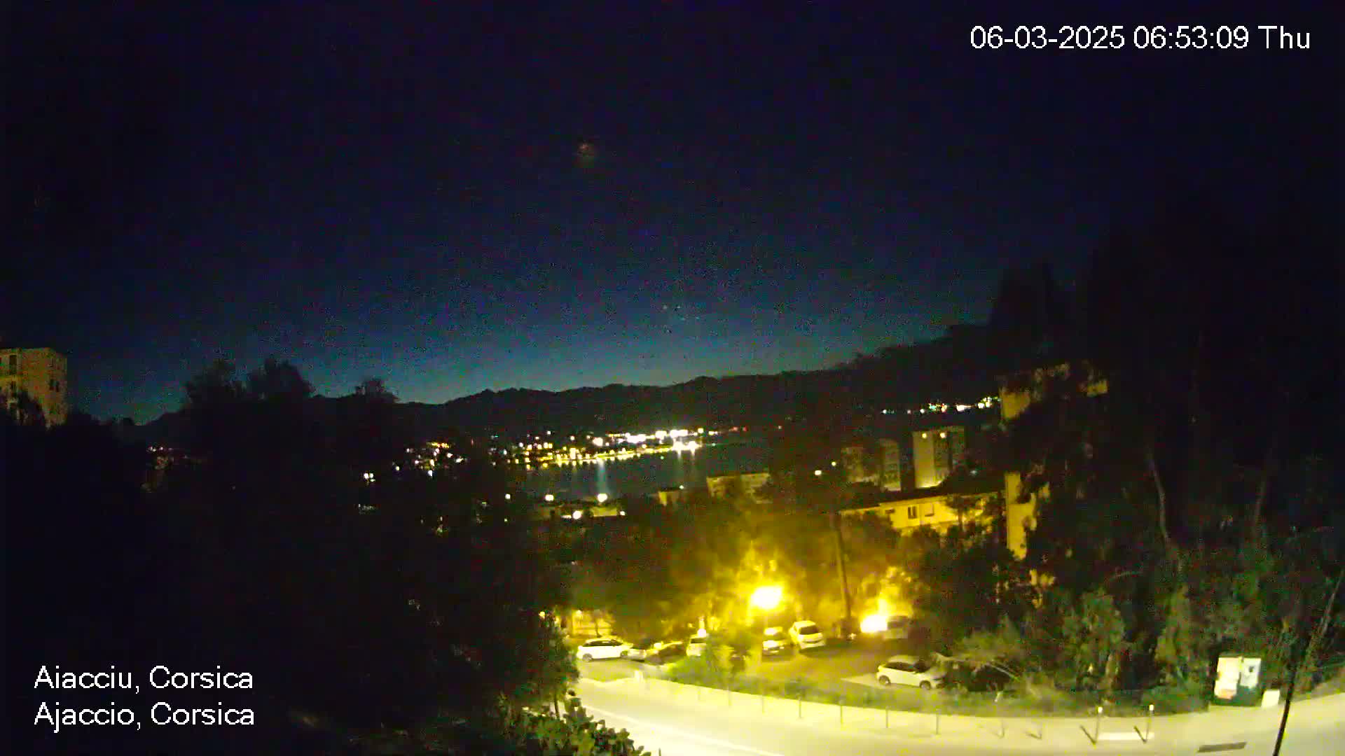 A nighttime, mostly clear view overlooking a city and body of water illuminated with lights, nestled against dark mountains.