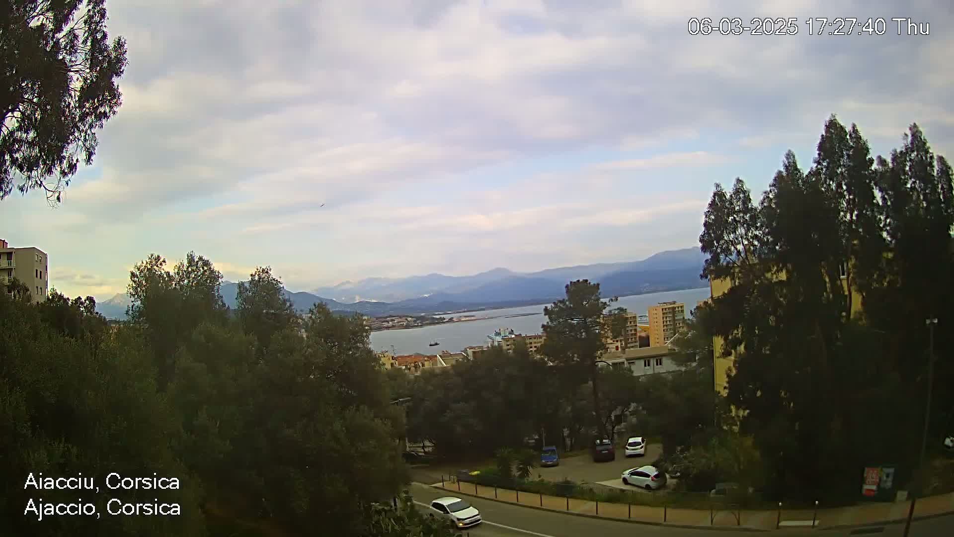 A partly cloudy day reveals a coastal view of buildings, trees, a body of water, and distant mountains.