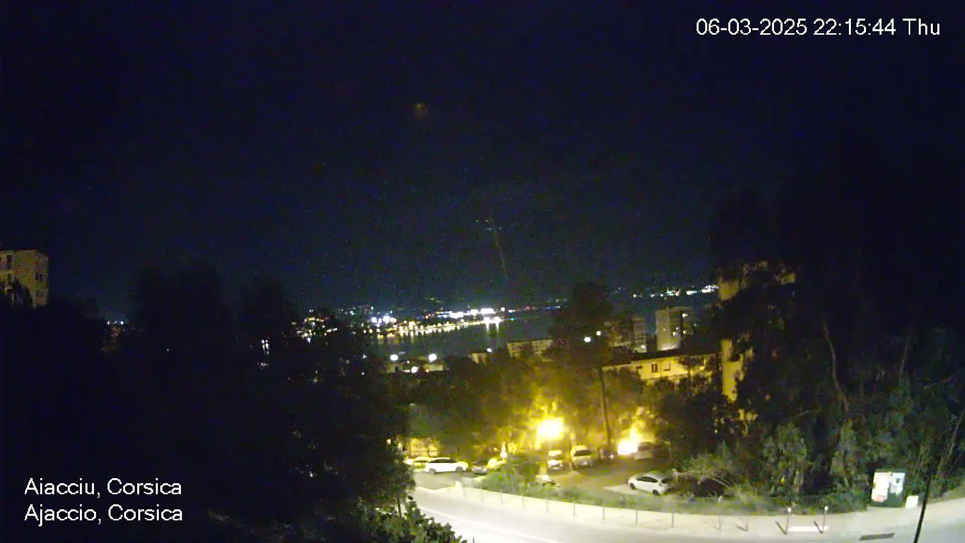 A nighttime view of a coastal city, with dimly lit buildings, roads, and parked cars, overlooking a body of water with lights visible in the distance under a clear, dark sky.