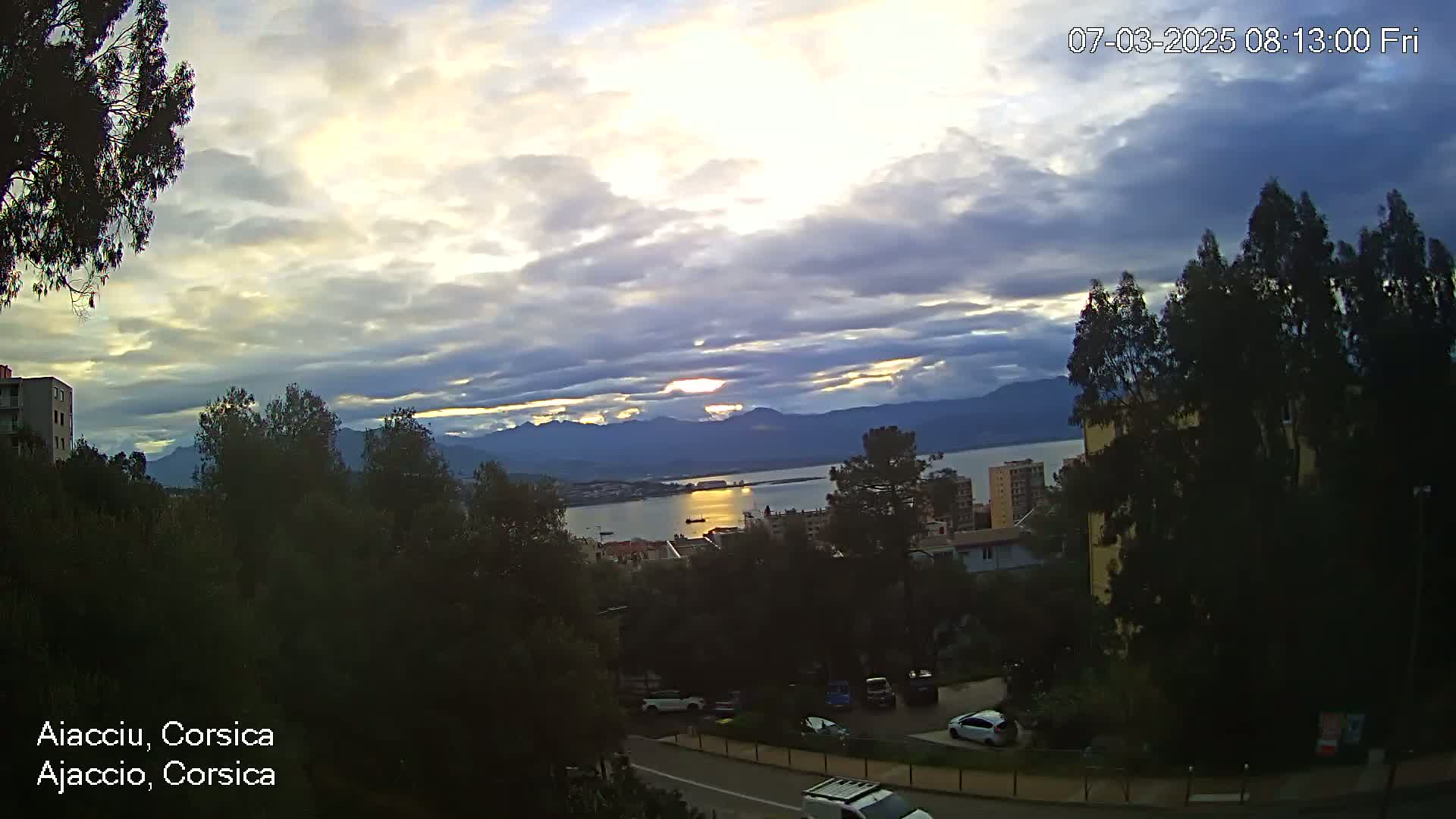 A partly cloudy sunrise over a calm body of water, with mountains in the distance and a city nestled among trees in the foreground.