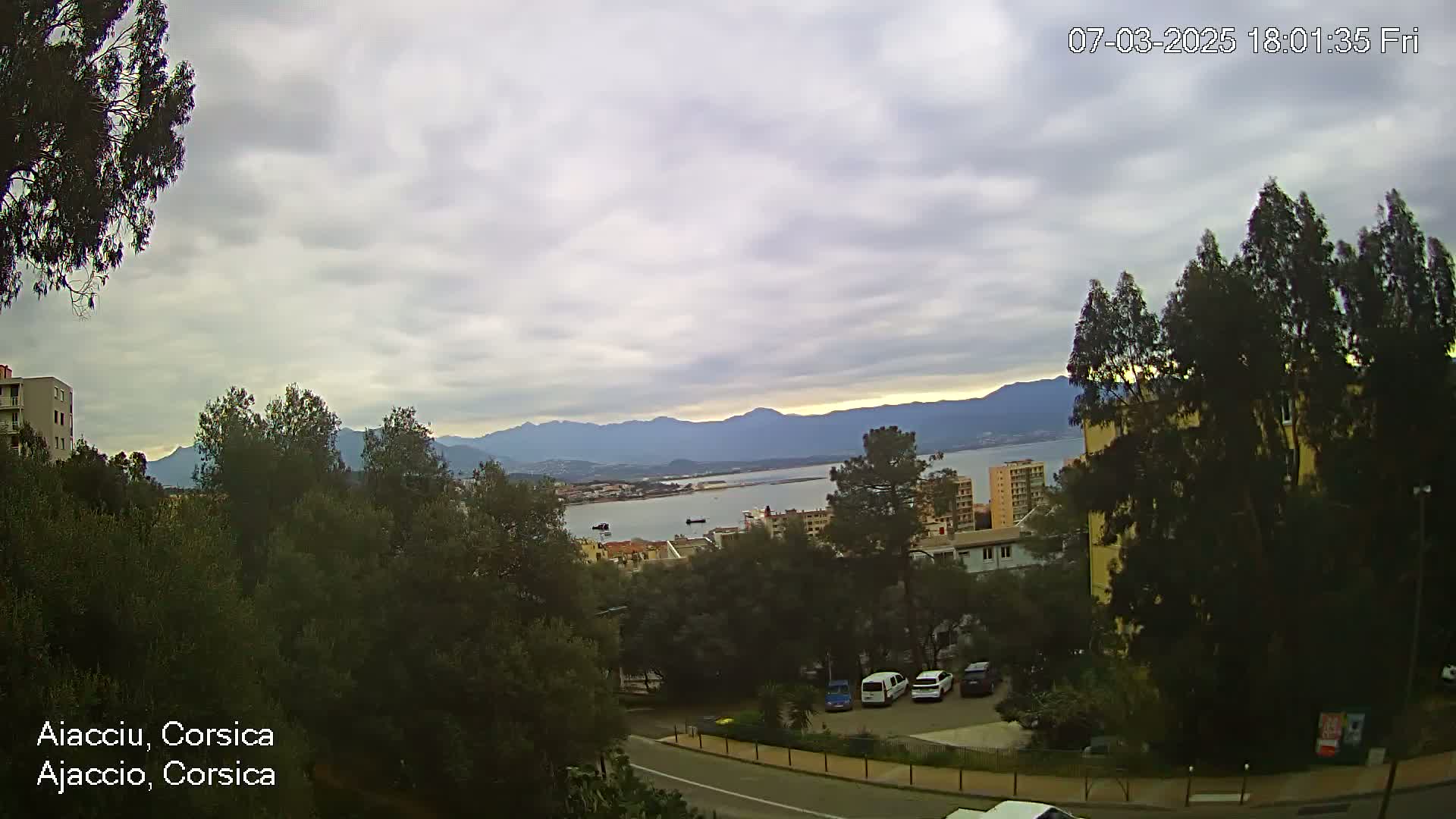 A mostly cloudy day reveals a coastal view of a city nestled among green trees and hills, with a body of water and distant mountains visible in the background.
