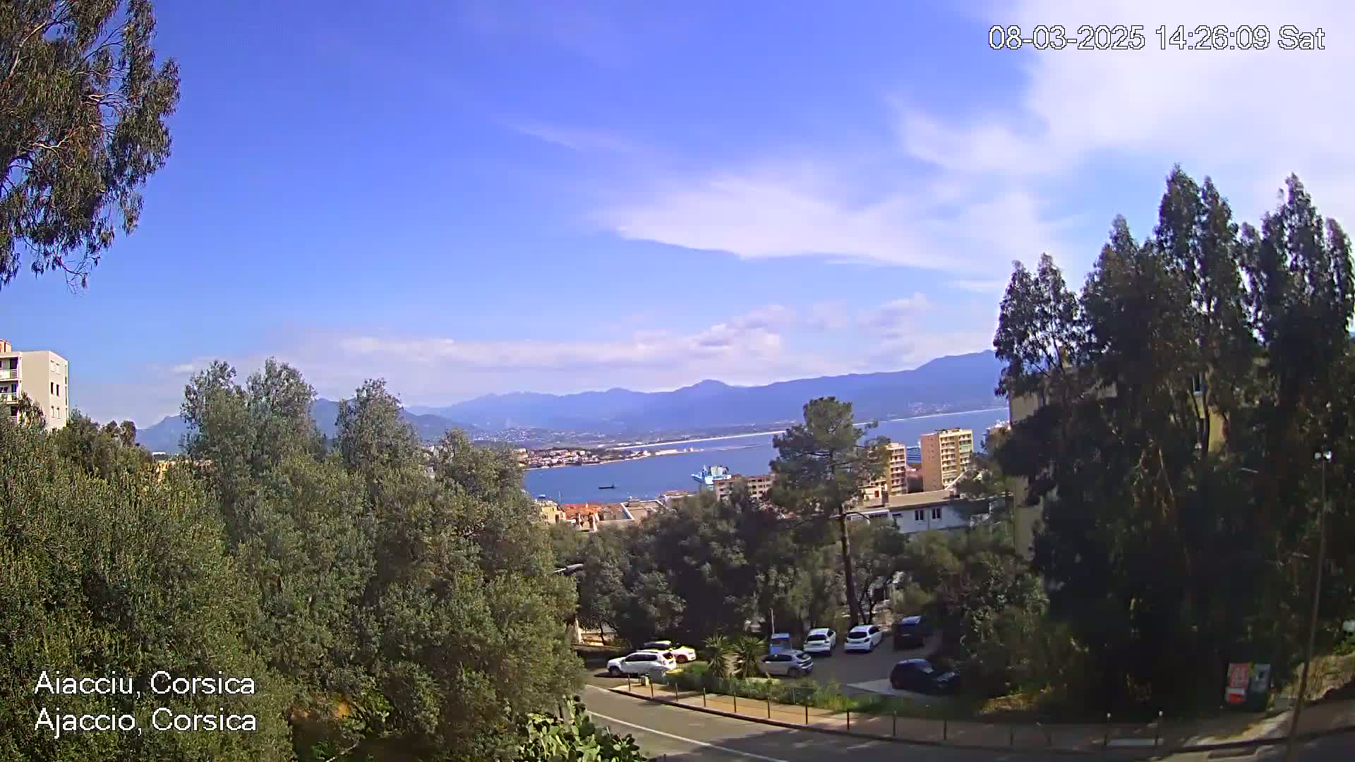 A partly sunny day reveals a coastal town nestled amongst green trees and hills, overlooking a calm bay and distant mountains.