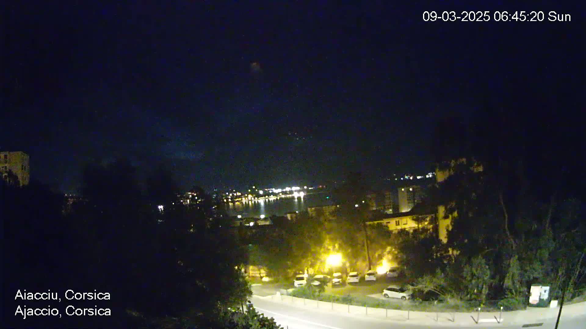 A nighttime view of a coastal town with a body of water and city lights visible in the distance under a clear, dark sky.