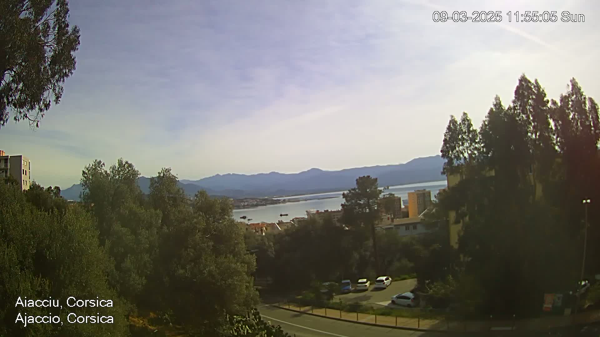 A partly cloudy day reveals a coastal town nestled among green trees and hills, overlooking a calm body of water and distant mountains.