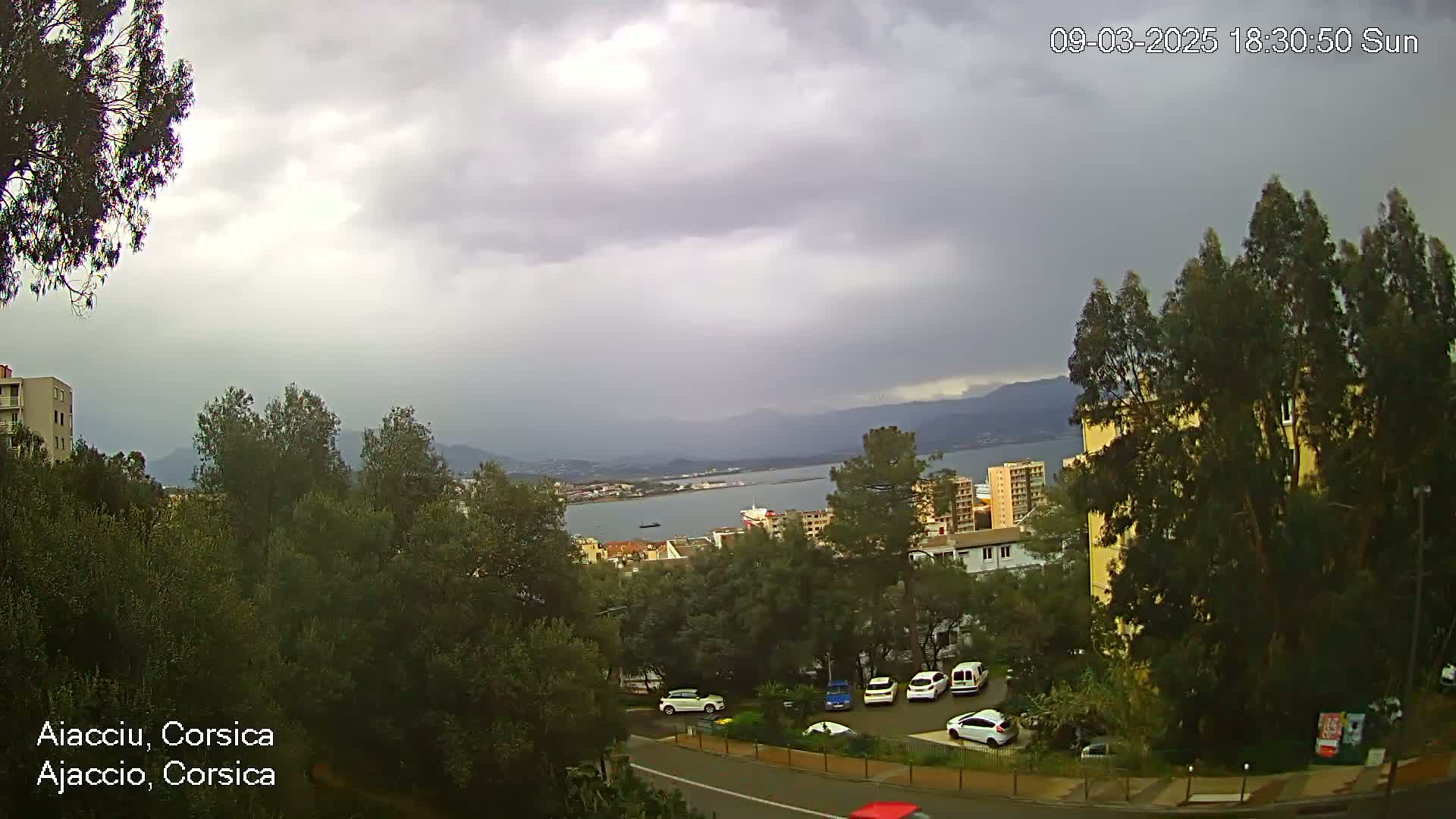A cloudy, overcast day reveals a coastal city scene with buildings, green trees, parked cars, and a body of water in the distance, mountains visible on the horizon.