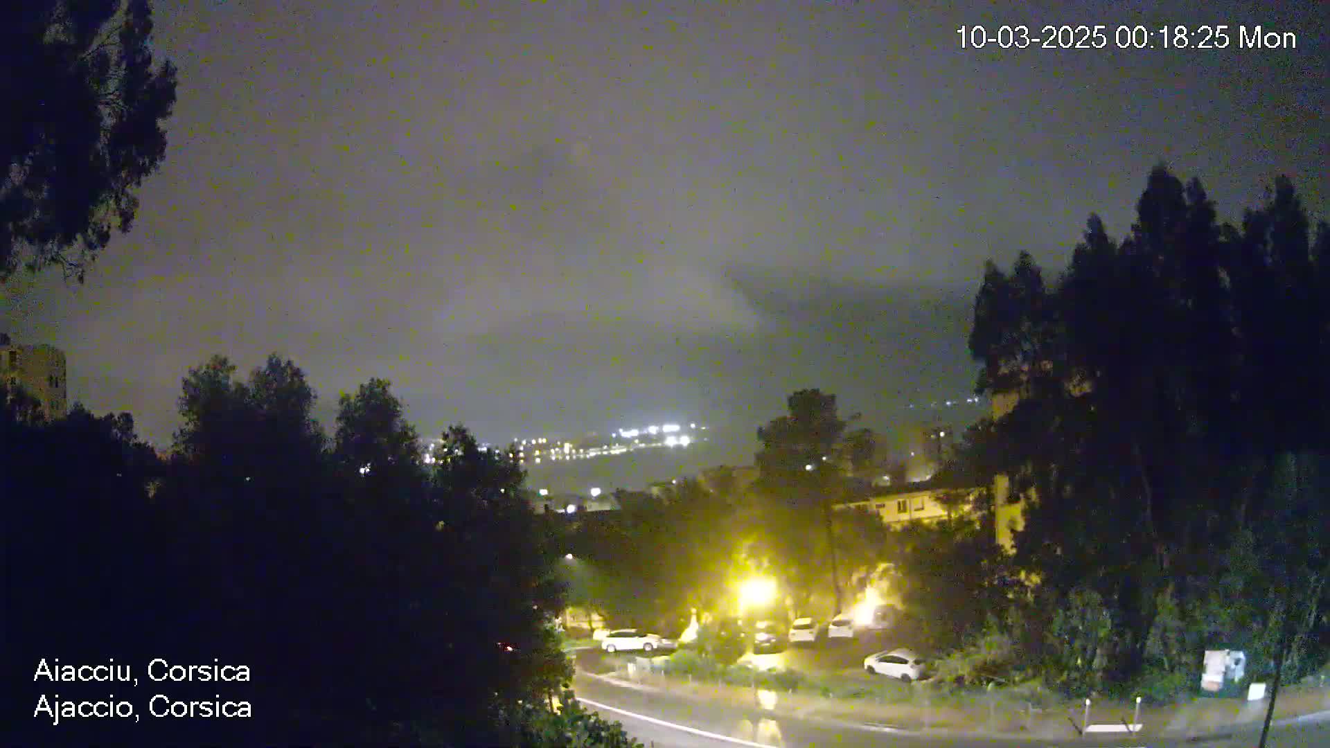 A nighttime view of a city shrouded in dark clouds and light rain, with a few cars parked along a wet road, and city lights faintly visible in the distance.