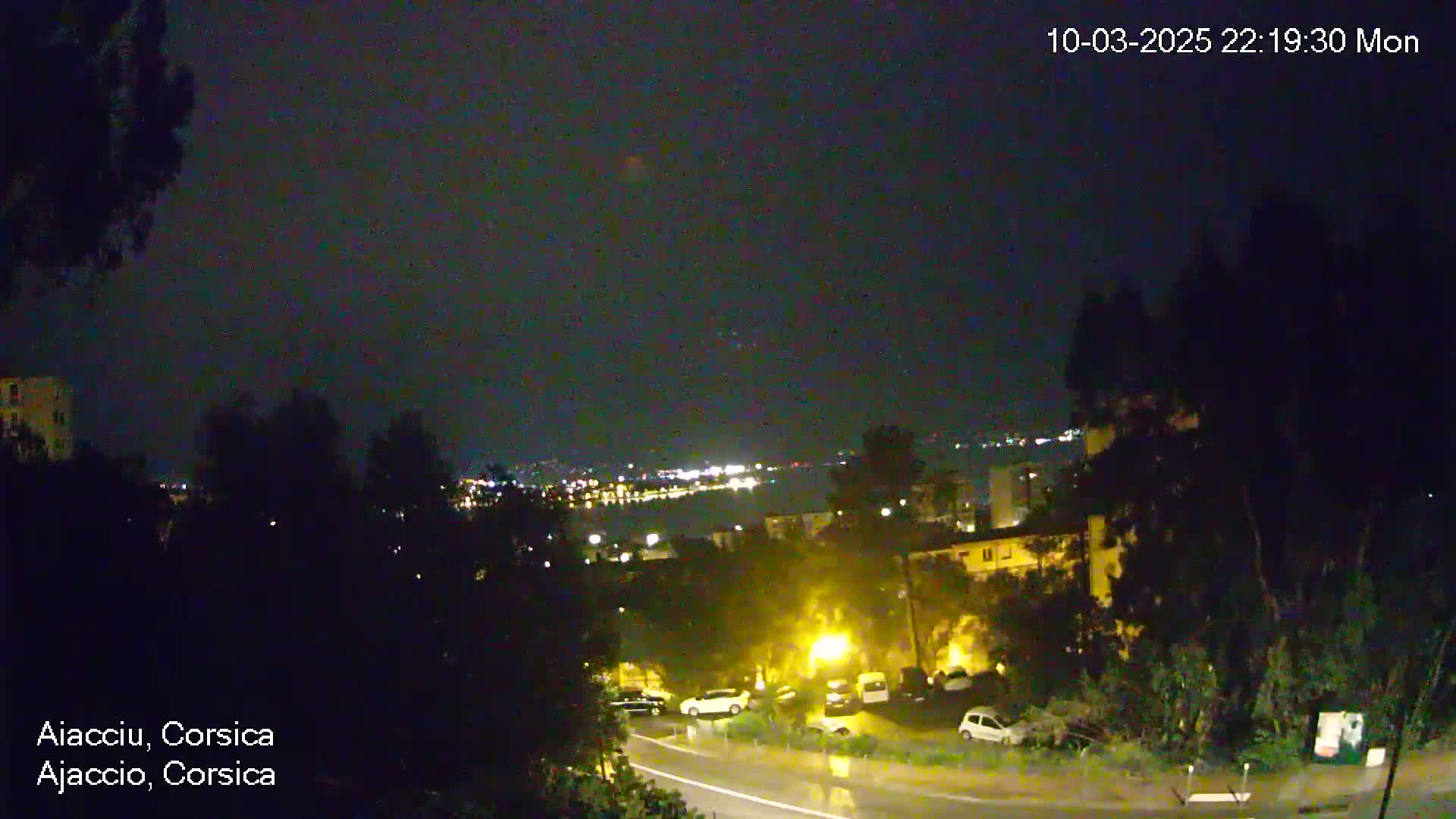 A nighttime view of a city overlooking a body of water with lights visible in the distance, under a dark, possibly overcast sky, with some wet roads visible in the foreground.