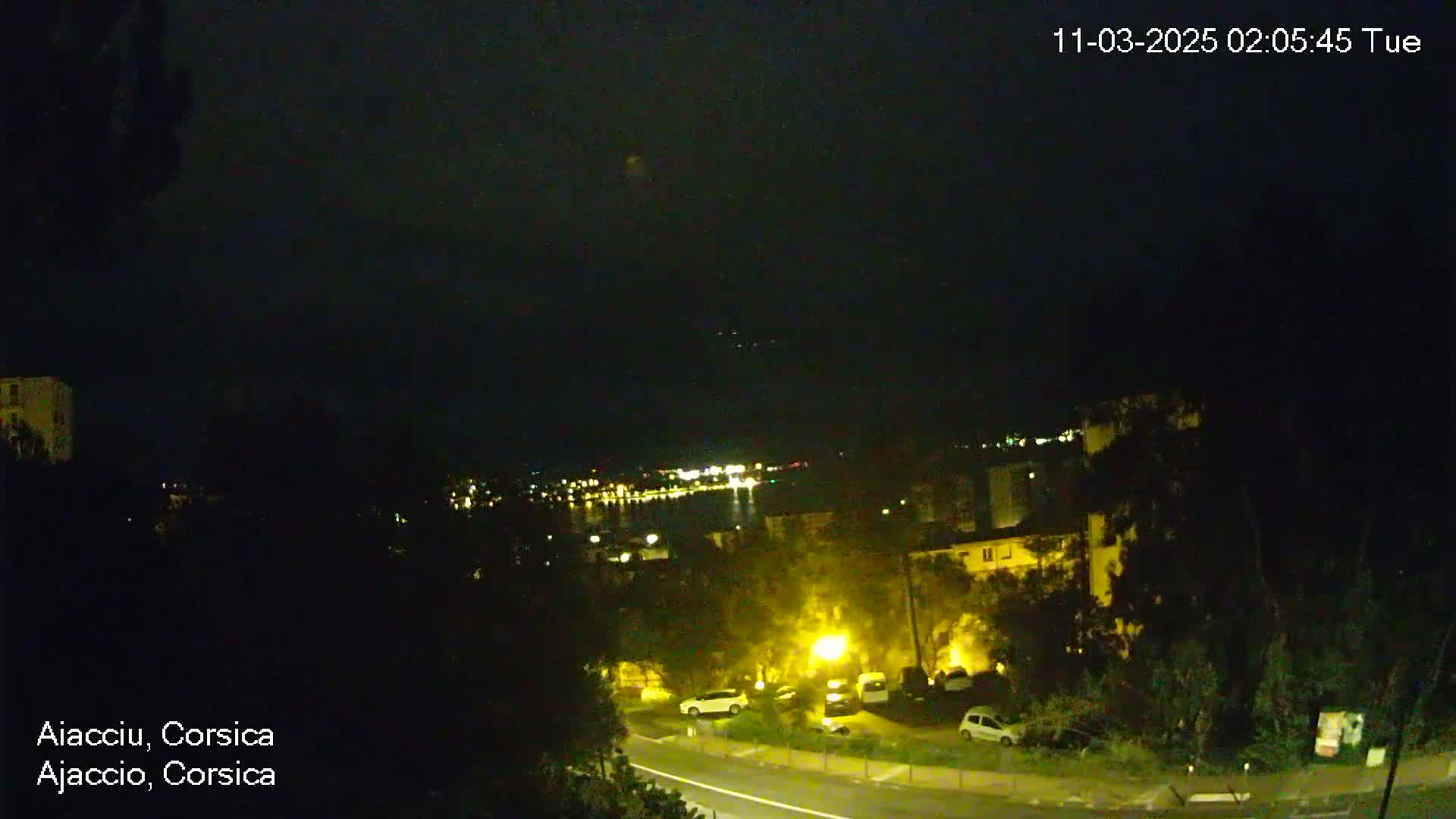 A nighttime, high-angle view shows a coastal town with dimly lit buildings, parked cars, and a dark body of water with faint lights in the distance.
