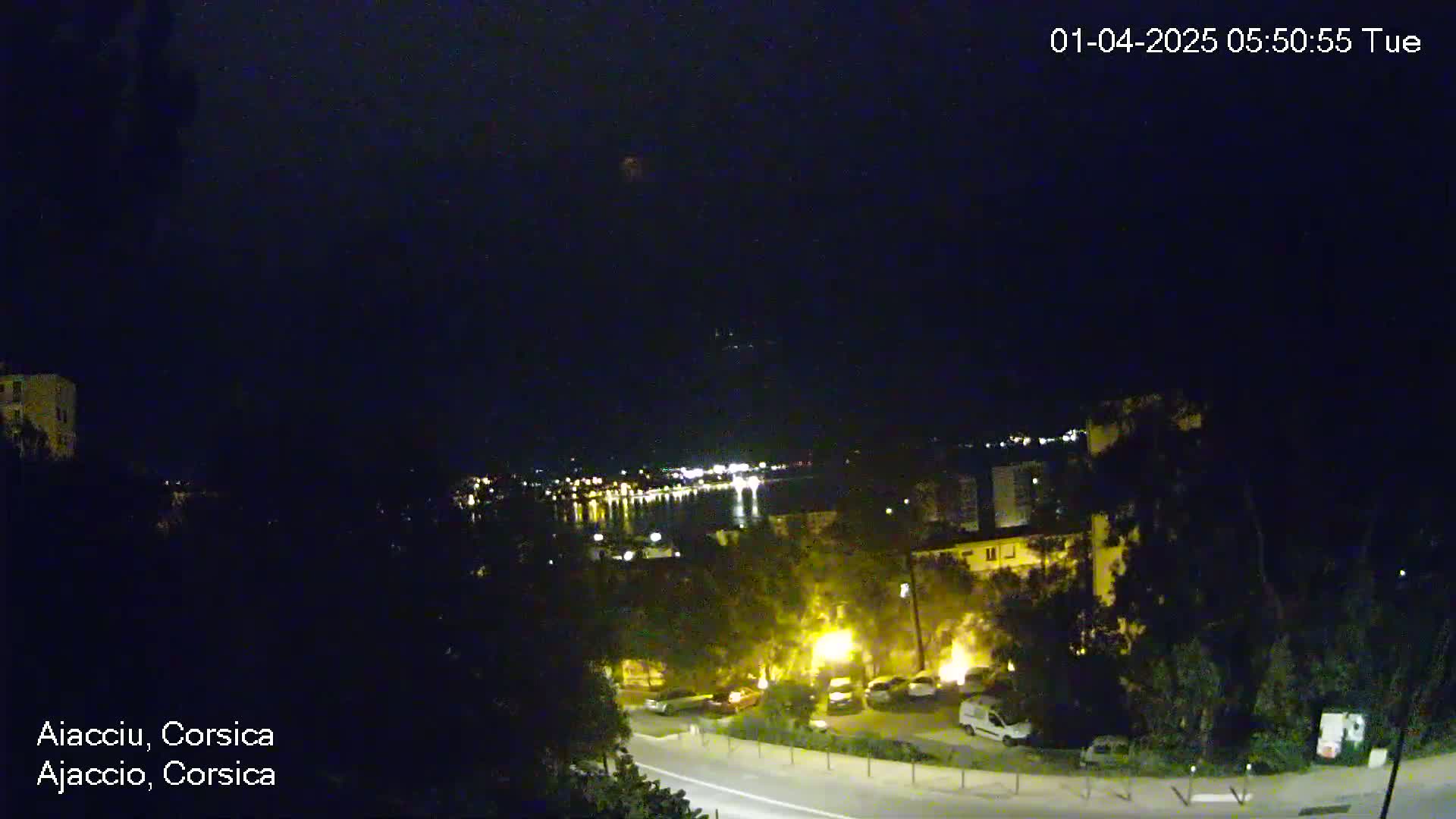 A nighttime, overhead view of a coastal town shows a road with parked cars and buildings illuminated by streetlights, overlooking a dark body of water with distant lights across the shore.