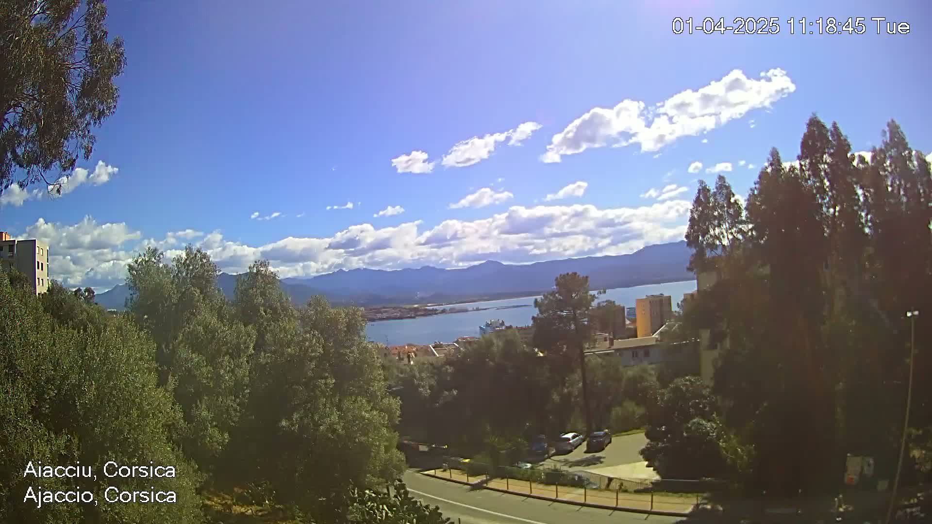 A partly cloudy sky overlooks a coastal town nestled amongst green trees and mountains, with a body of water visible in the distance.