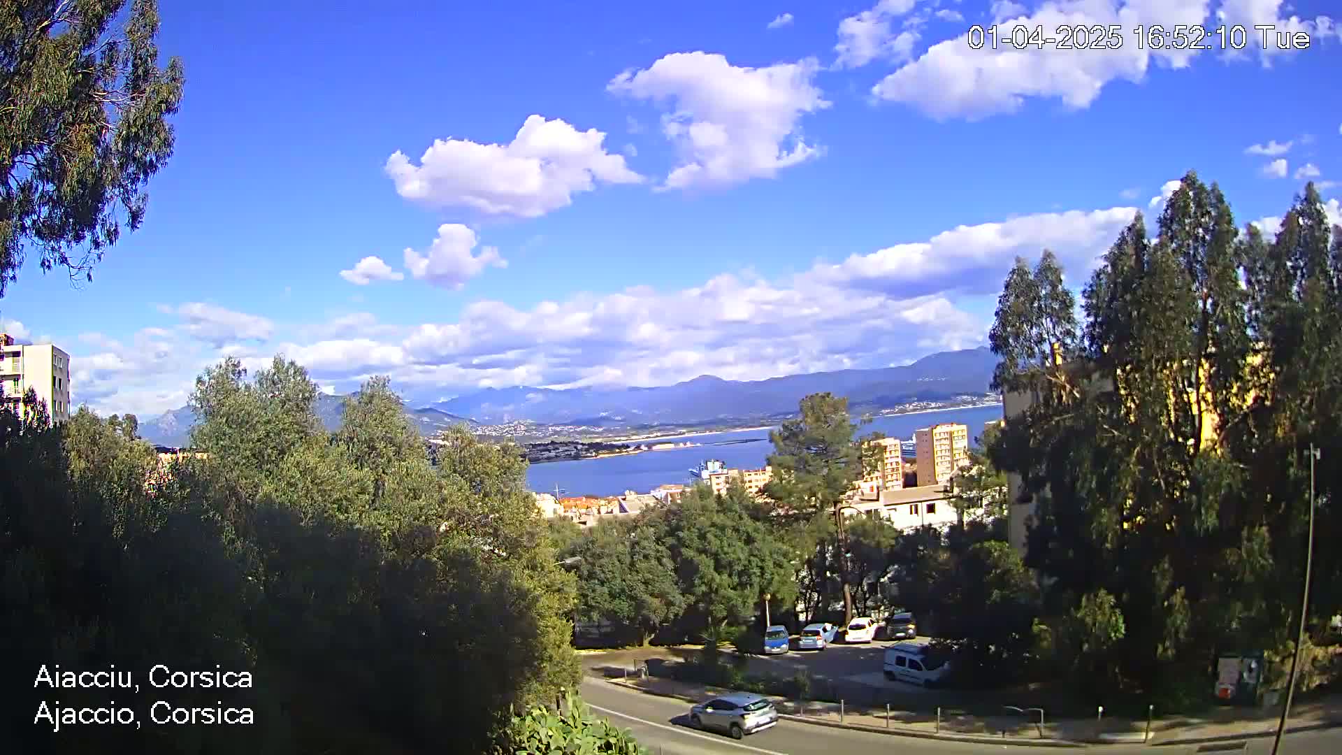 A partly cloudy blue sky overlooks a coastal city with green trees, buildings, and parked cars, mountains visible in the distance.