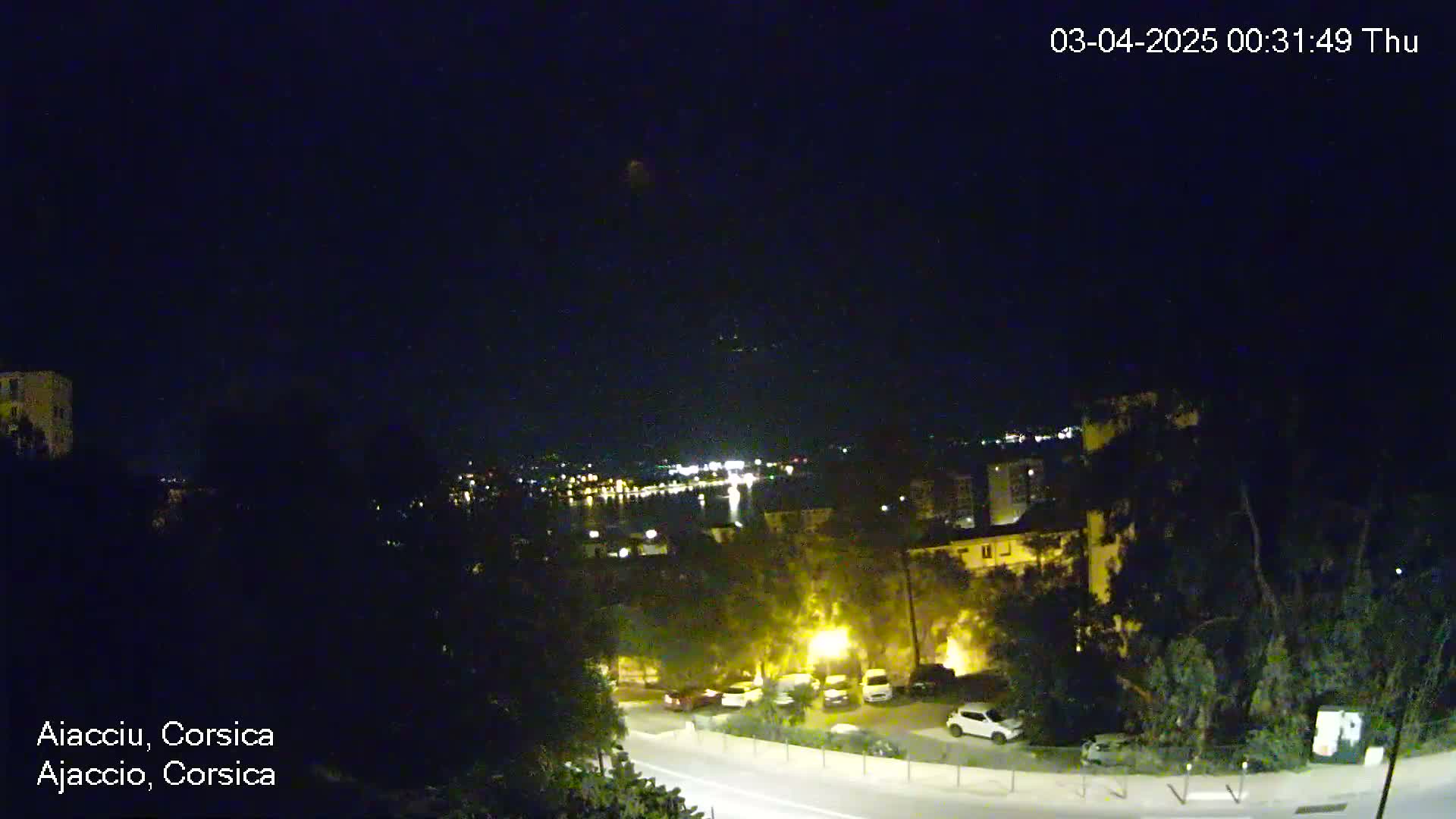 A nighttime, overhead view of a coastal town with a road curving through it, overlooking a body of water with lights visible in the distance under a clear, dark sky.