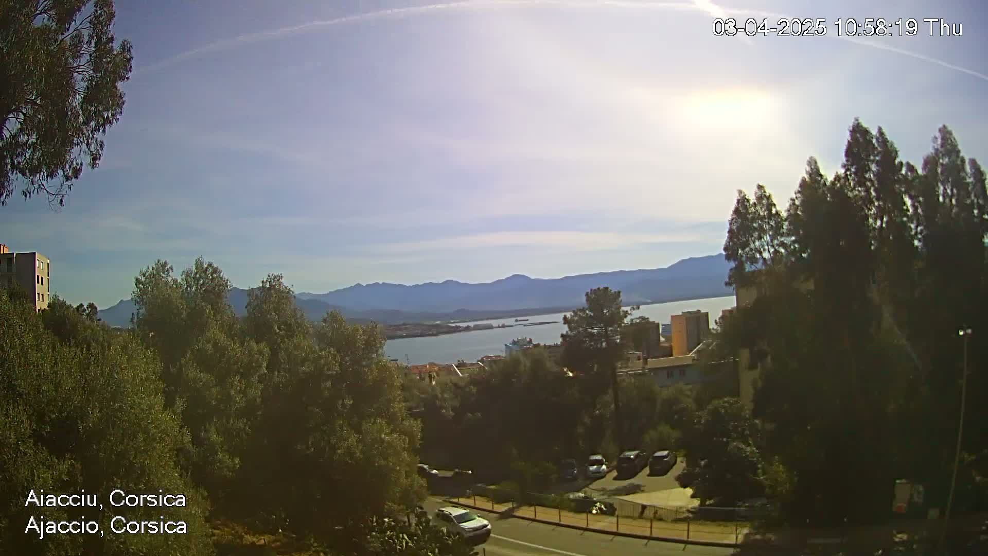 A sunny day reveals a panoramic view of a coastal town nestled among green hills, with a calm body of water and distant mountains visible in the background.