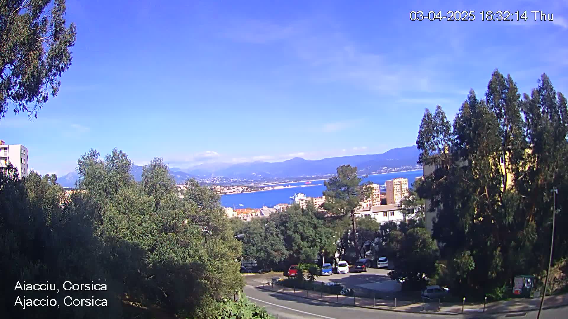 A sunny, clear day overlooks a coastal town nestled amongst lush green trees and mountains, with a body of water visible in the distance and cars parked in a lot nearby.