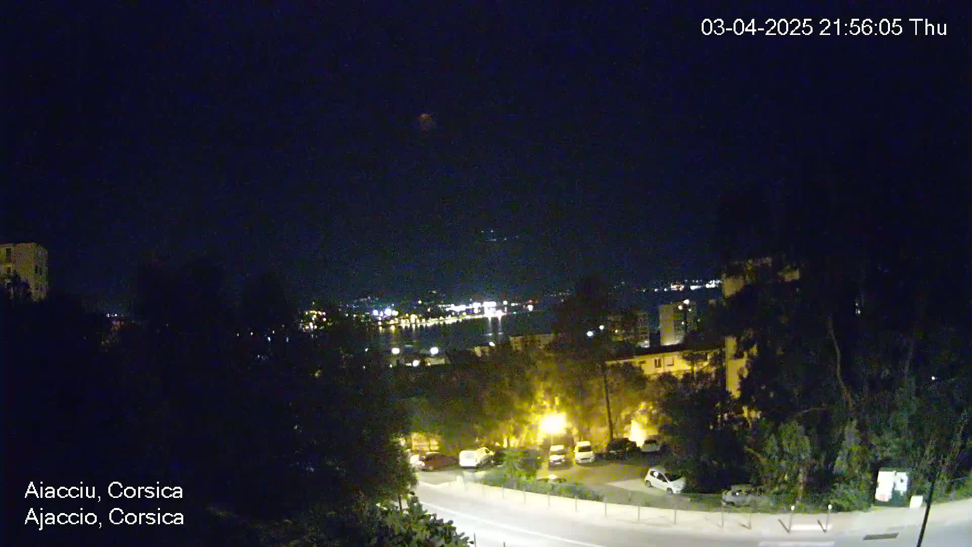 A nighttime view of a coastal town, with dimly lit buildings, parked cars, and a road visible under a clear, dark sky.
