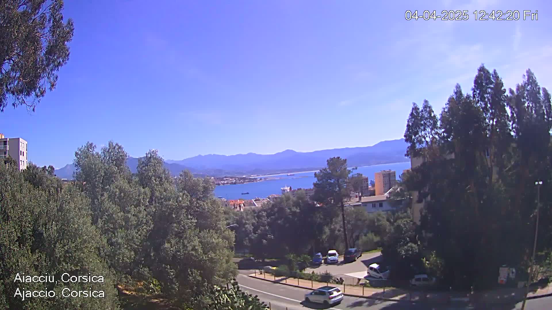 A clear, sunny day reveals a coastal town nestled among green trees and hills, with a calm body of water and distant mountains visible in the background.