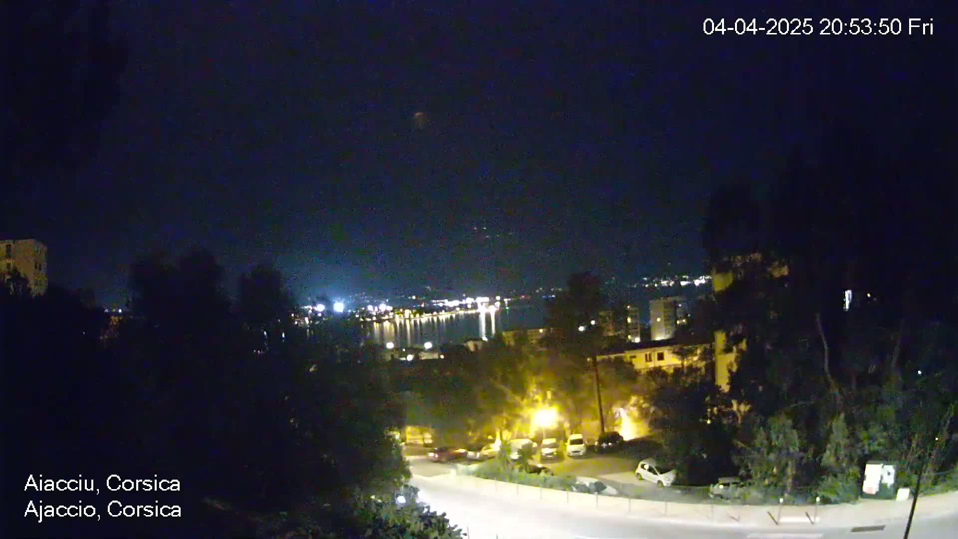 A nighttime, high-angle view of a city by the water, showing a road curving through a residential area with parked cars and streetlights under a clear, dark sky.