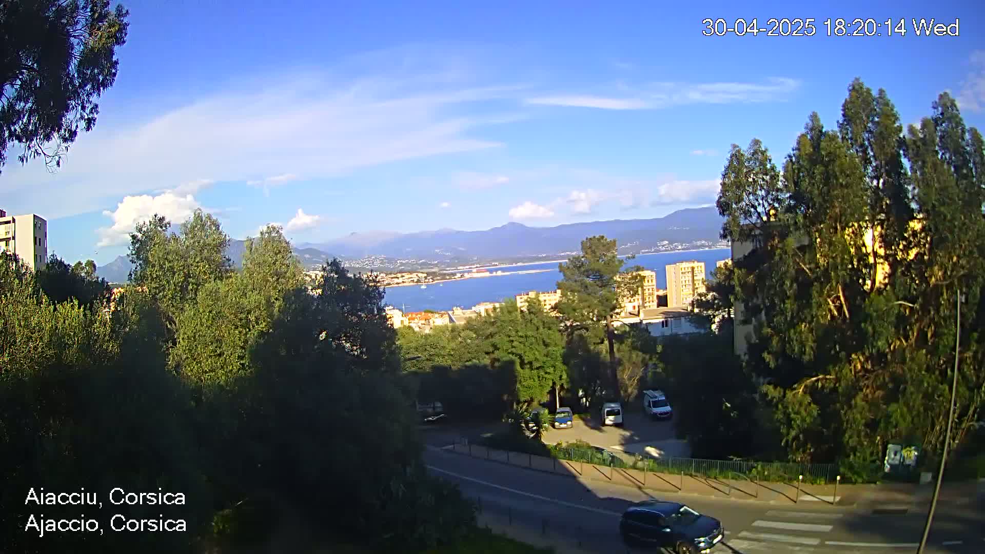 A partly sunny view overlooking a coastal city, with a calm body of water, mountains in the distance, and trees and buildings in the foreground.