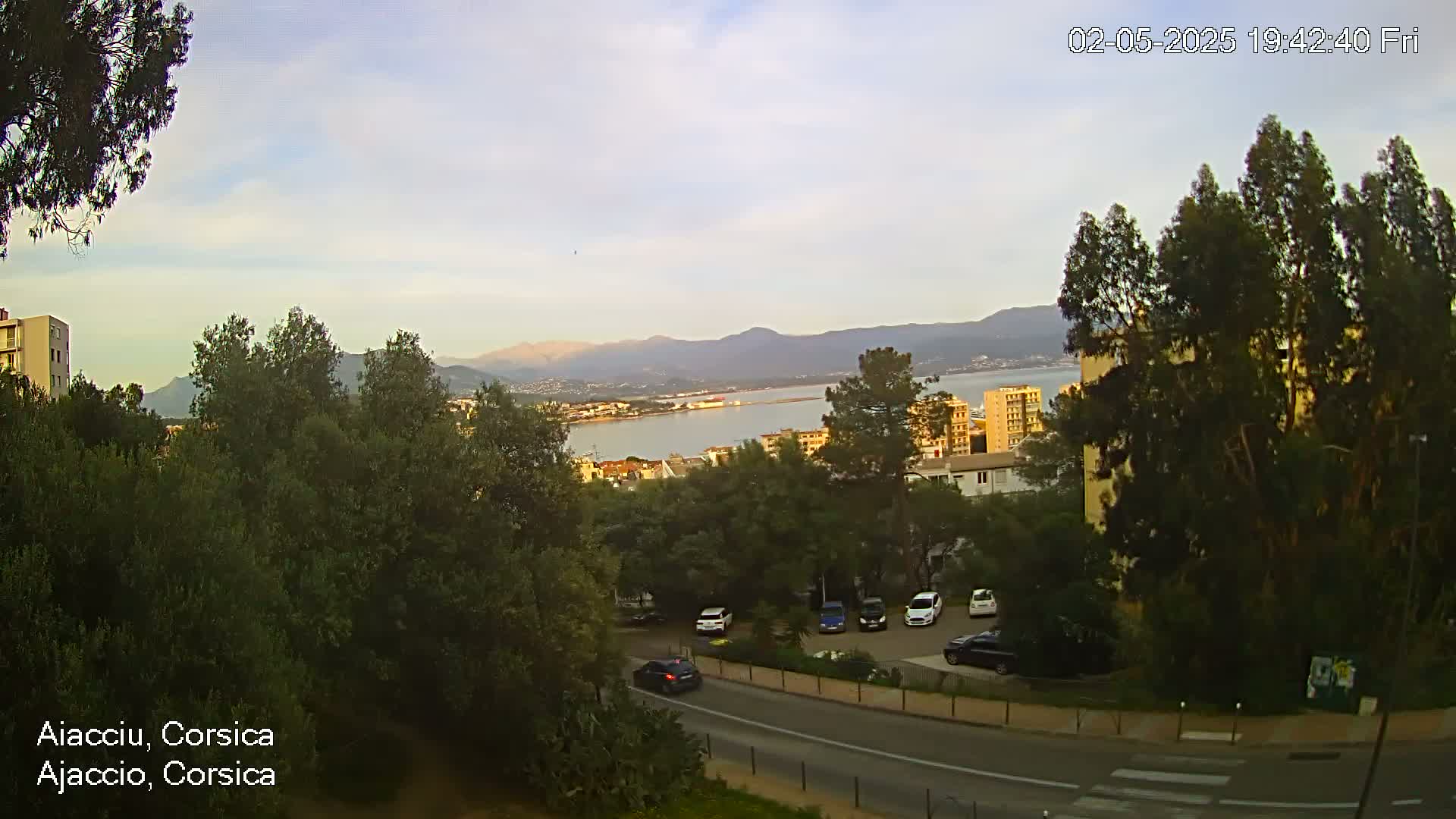 A coastal city scene under a partly cloudy sky shows a road winding through lush green vegetation, with parked cars and distant buildings overlooking a calm body of water and mountains in the background.