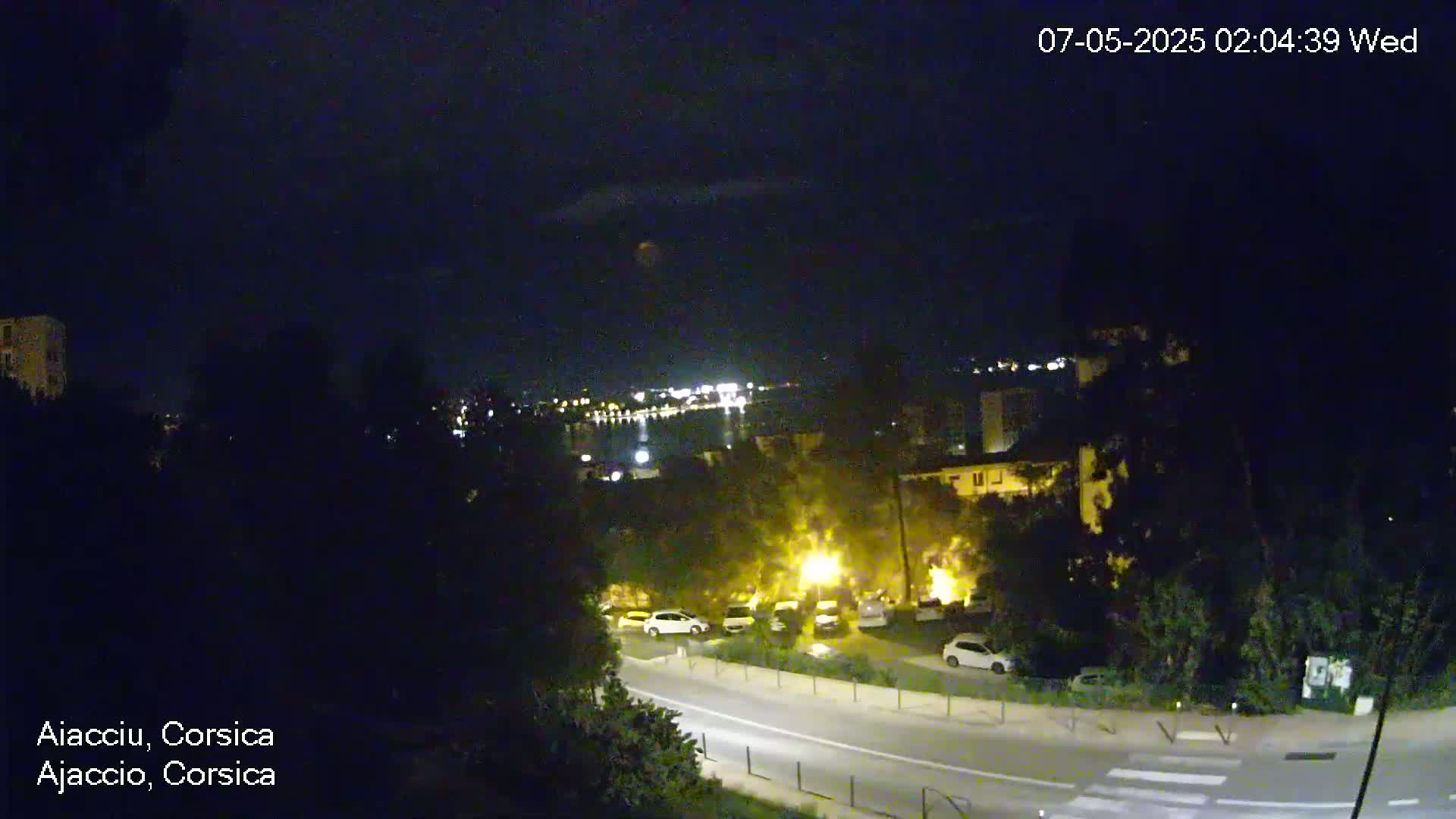 A nighttime view of a town with a road, parked cars, and buildings, overlooking a body of water with distant lights under a clear, dark sky.