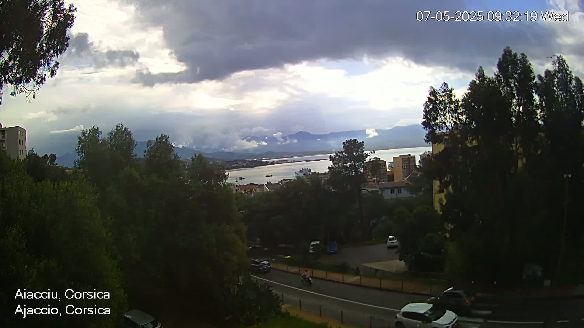 A partly cloudy day overlooks a body of water, mountains in the distance, a road with cars, and a scattering of buildings nestled among green trees.