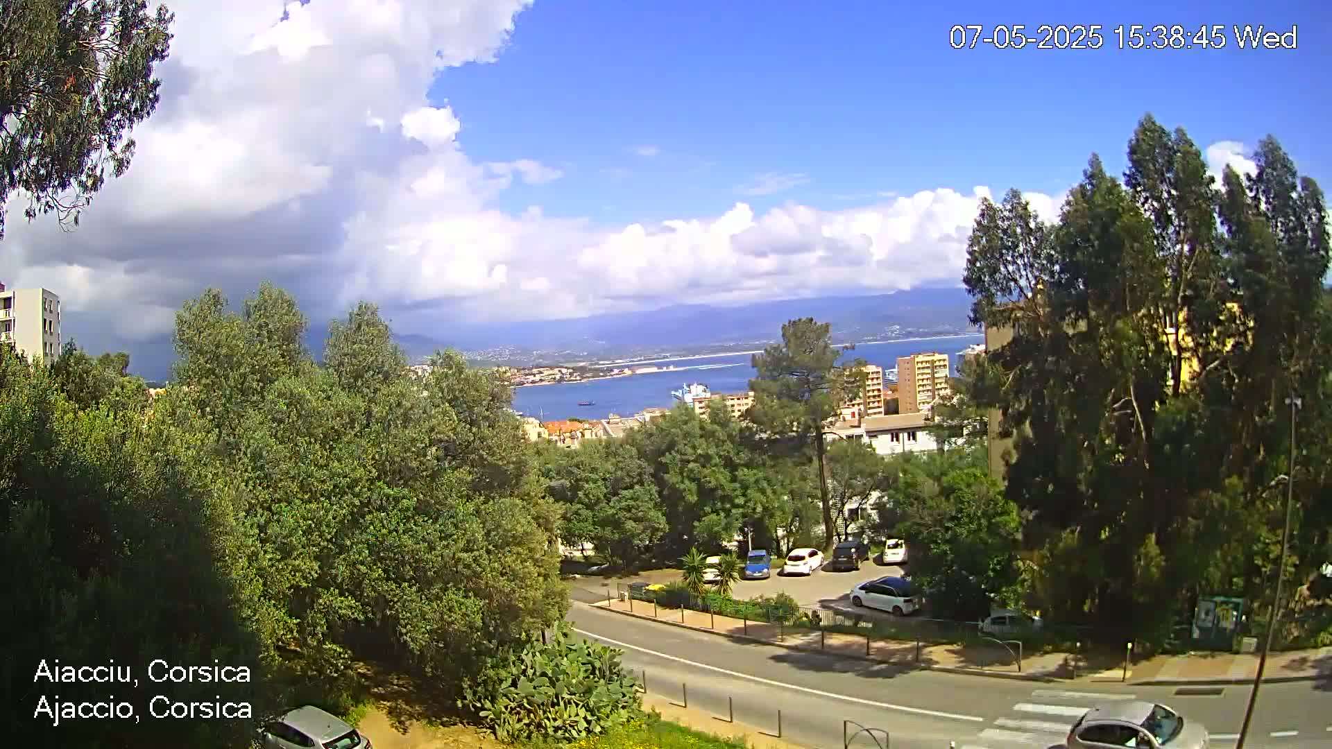 A partly cloudy day overlooks a coastal city with green trees, roads, parked cars, and a body of water in the distance.