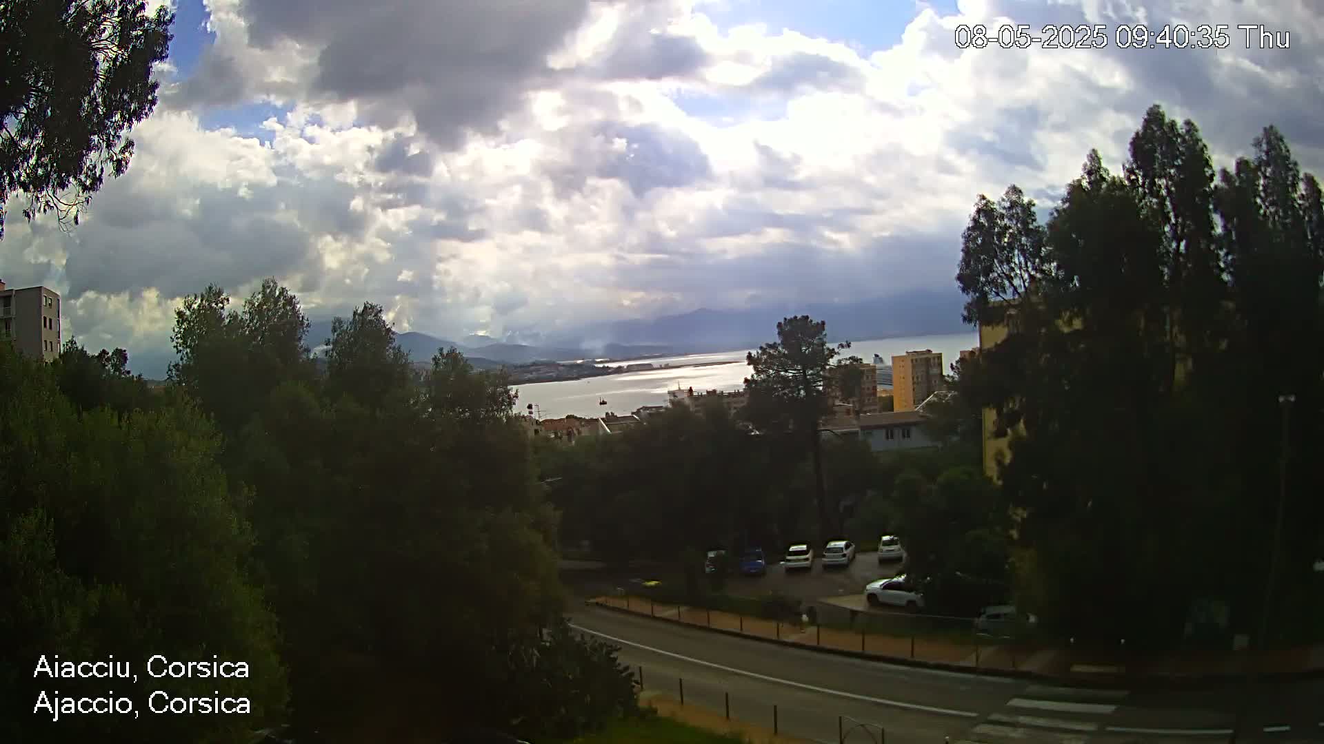 A partly cloudy sky overlooks a coastal town with a body of water and mountains in the distance, visible from an elevated vantage point.