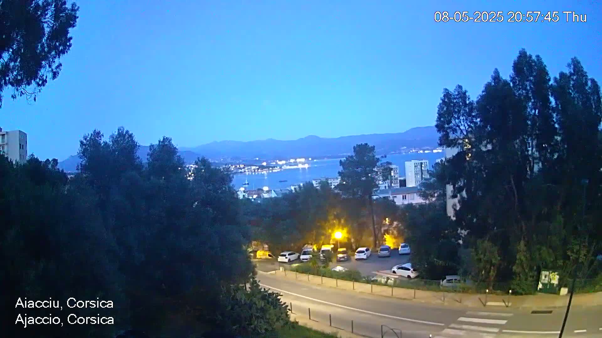 A twilight view of a coastal city with a calm body of water, mountains in the background, and a road with parked cars in the foreground.