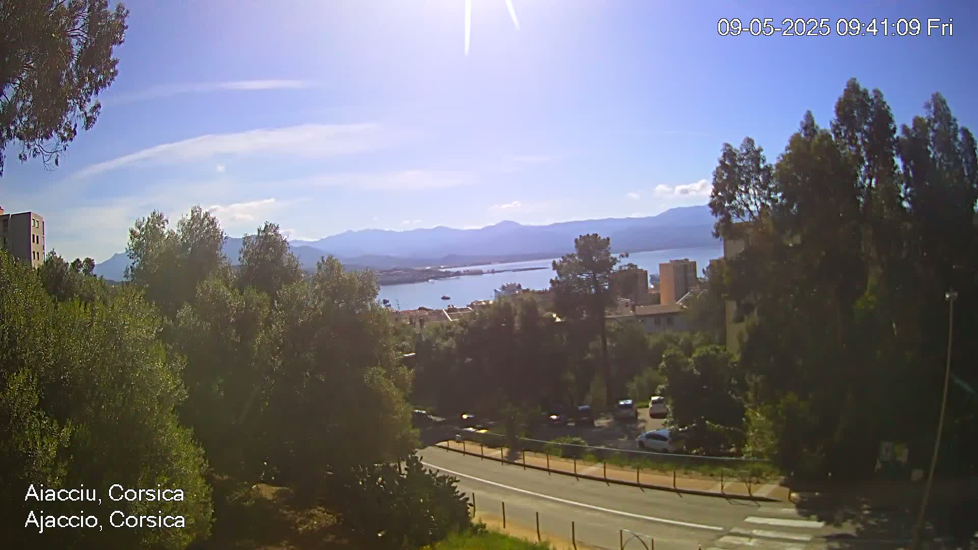 A sunny, partly cloudy day reveals a view of a city nestled among green trees, overlooking a calm body of water and distant mountains.