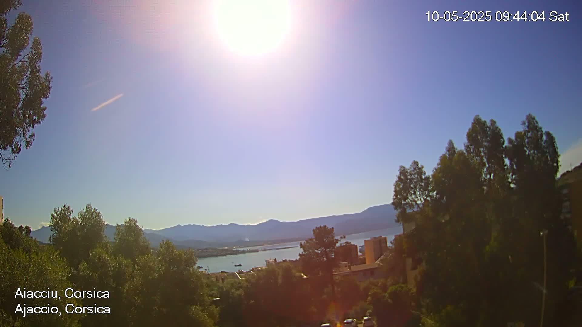 A sunny, clear day overlooks a town nestled among green trees and hills, with a body of water and distant mountains in the background.