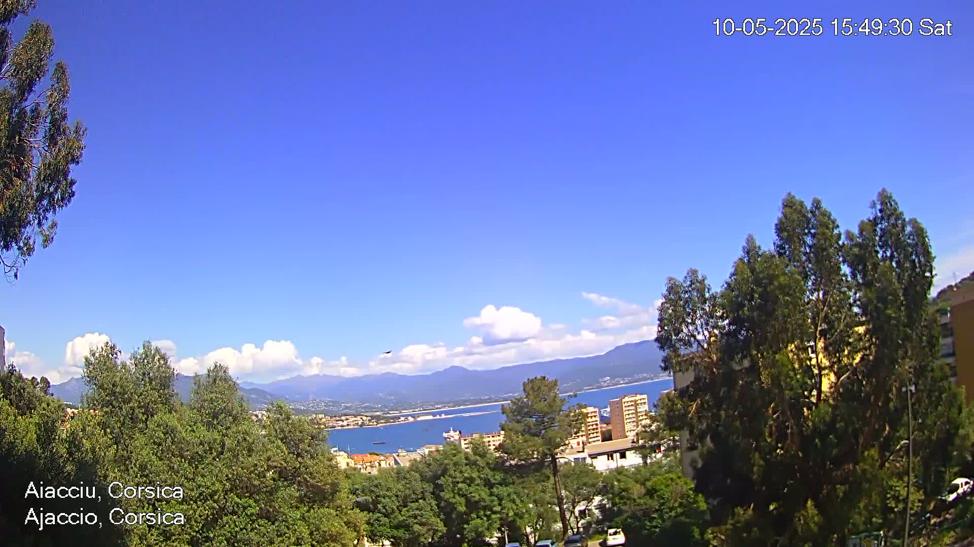 A partly sunny day reveals a coastal city scene with a blue body of water, mountains in the distance, and green trees in the foreground.