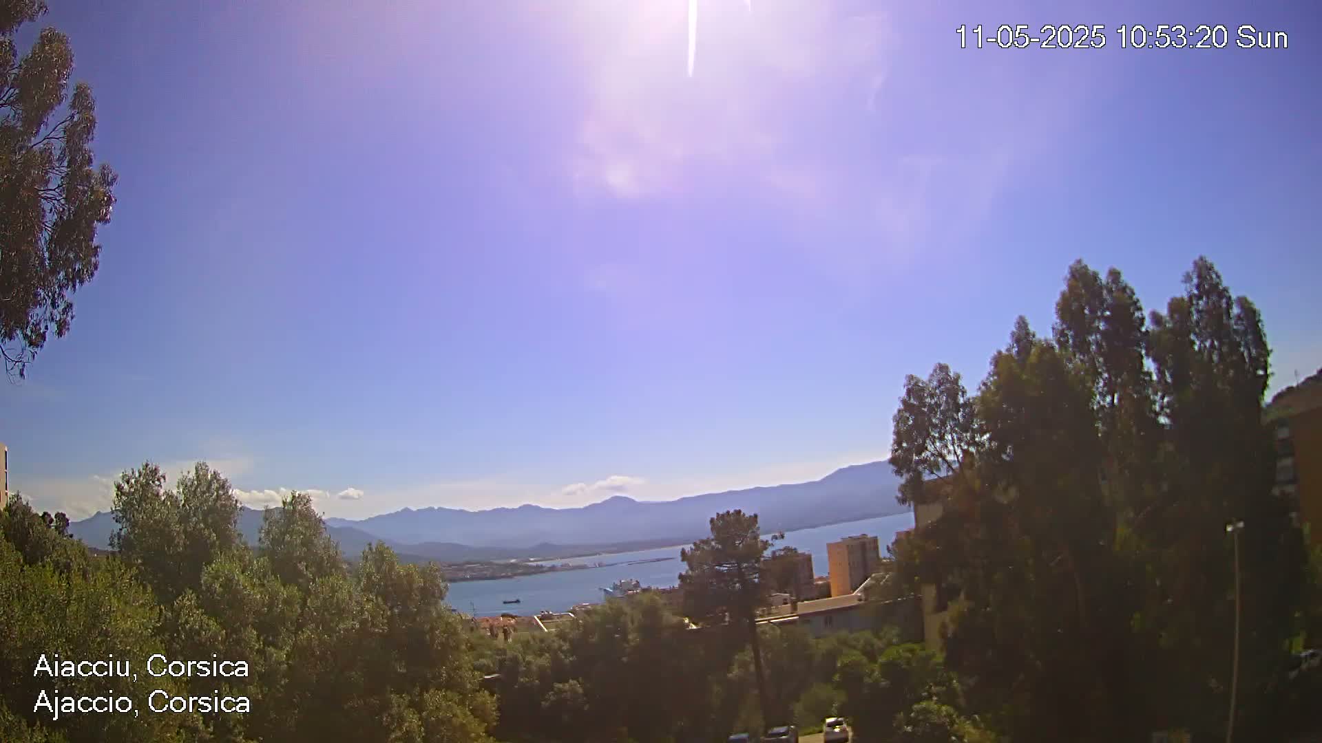A sunny, clear day overlooks a coastal town nestled among green trees and mountains, with a body of water visible in the distance.