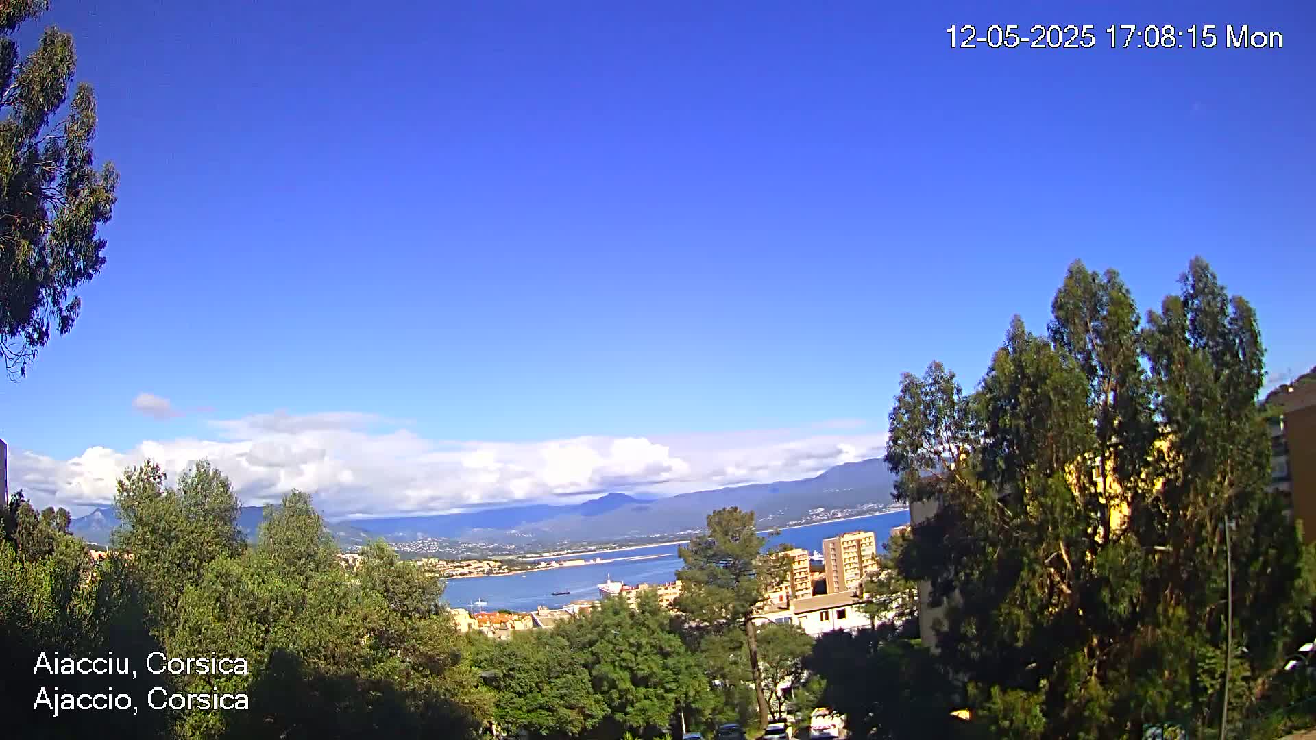A partly cloudy blue sky overlooks a coastal city with green trees and mountains in the distance.