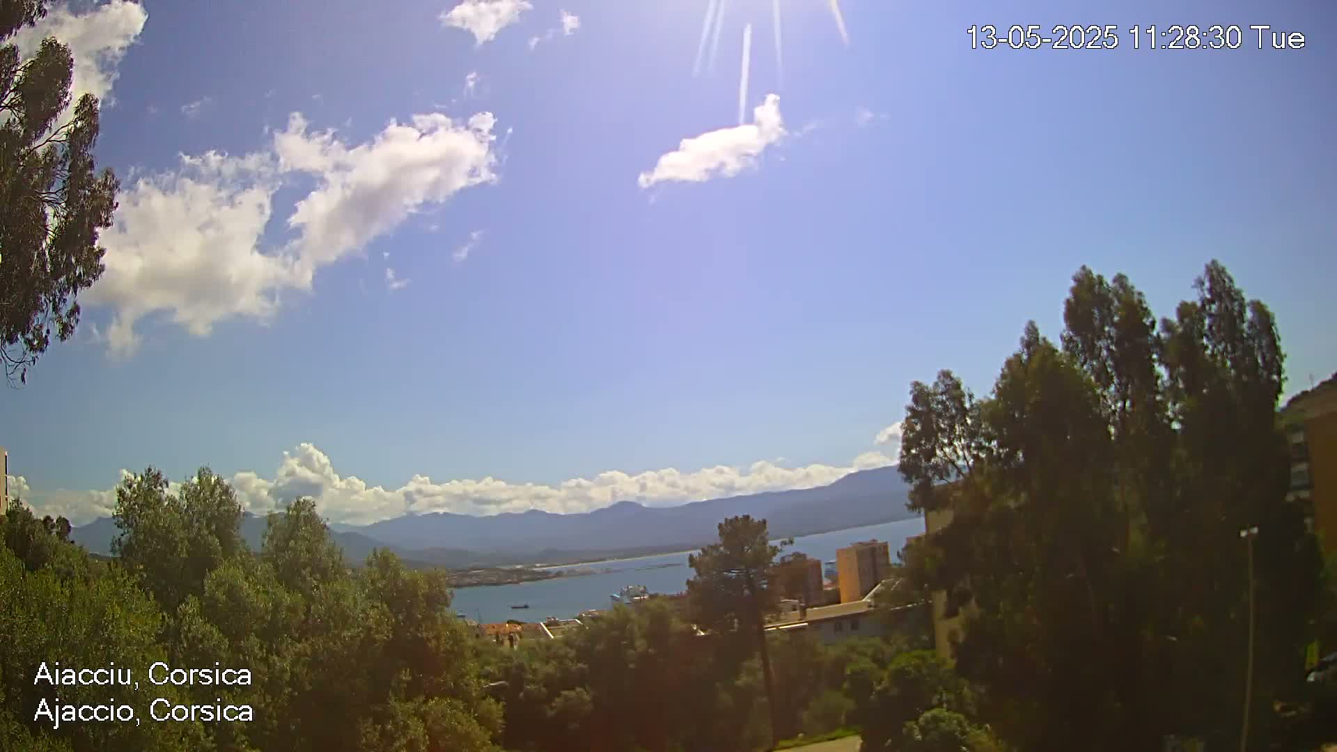 A sunny day with scattered clouds reveals a coastal view of buildings, trees, and distant mountains, overlooking a body of water.