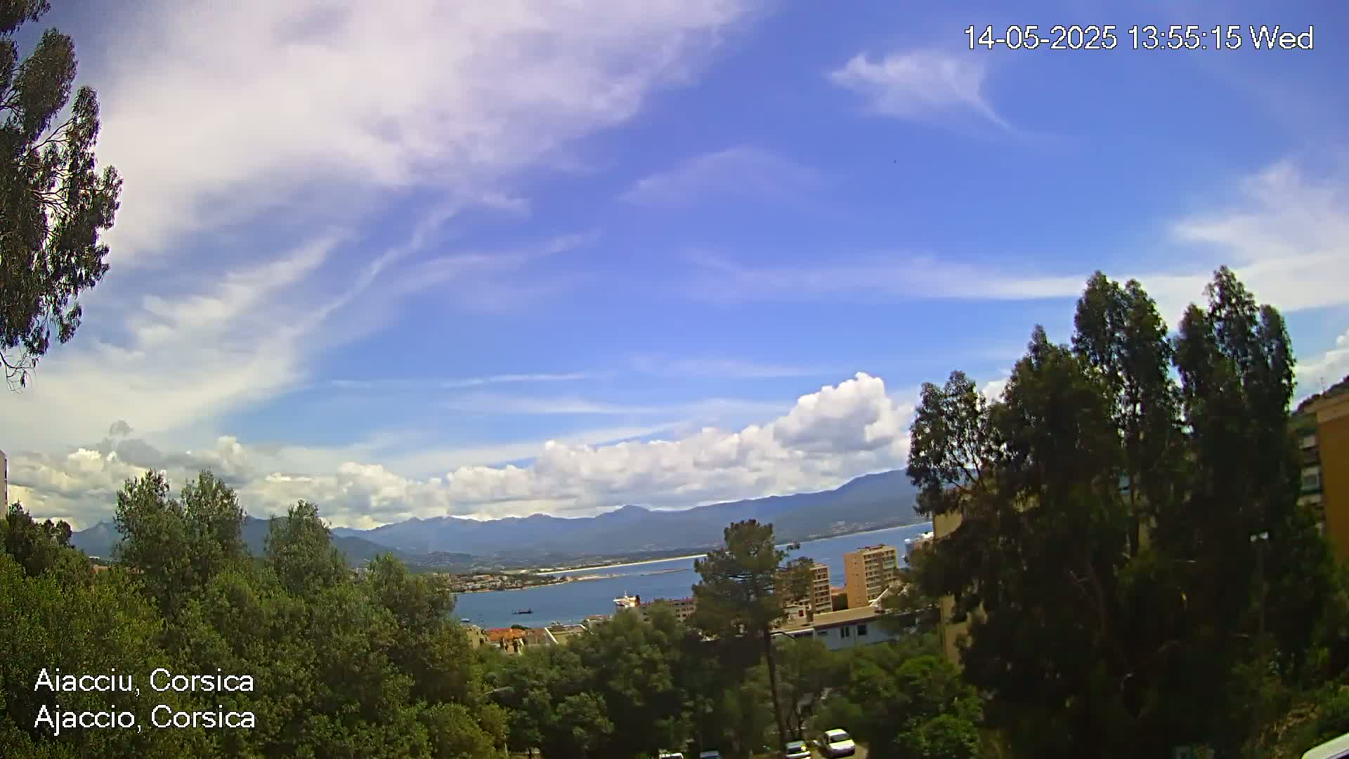 A partly cloudy blue sky overlooks a coastal town nestled among green trees and mountains, with a body of water visible in the distance.