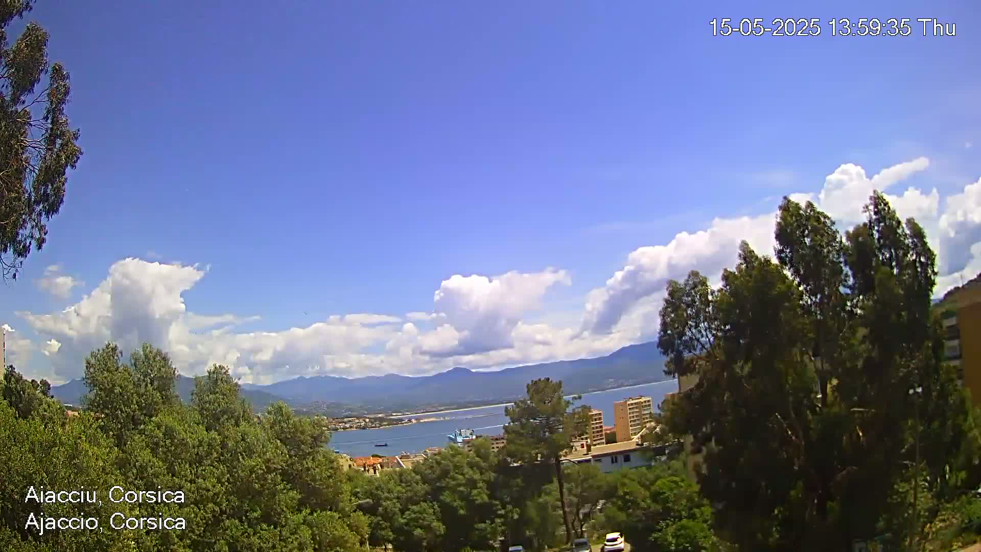 A partly cloudy sunny day overlooks a coastal town with a bay and mountains in the distance, viewed from a hilltop amidst lush green trees.