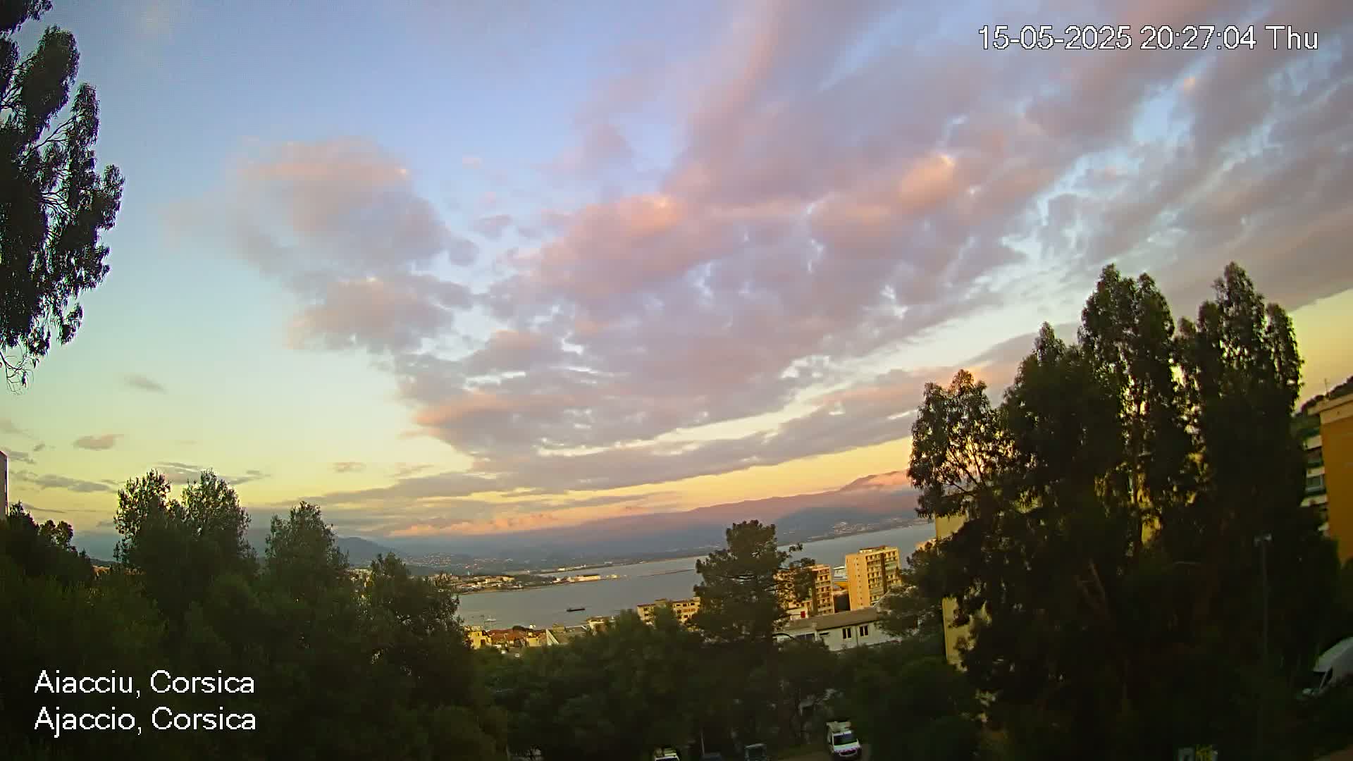 A partly cloudy sunset over a coastal city with numerous trees in the foreground.