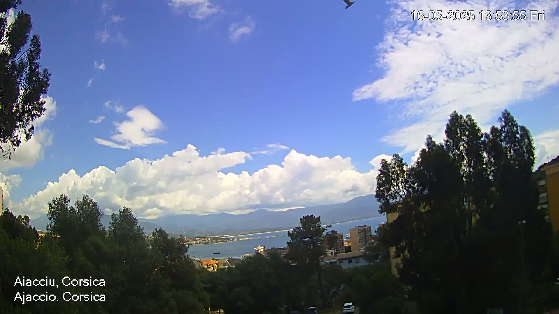 A partly cloudy sky with cumulus clouds over a coastal town nestled amongst green trees and mountains, under a mostly clear blue sky.