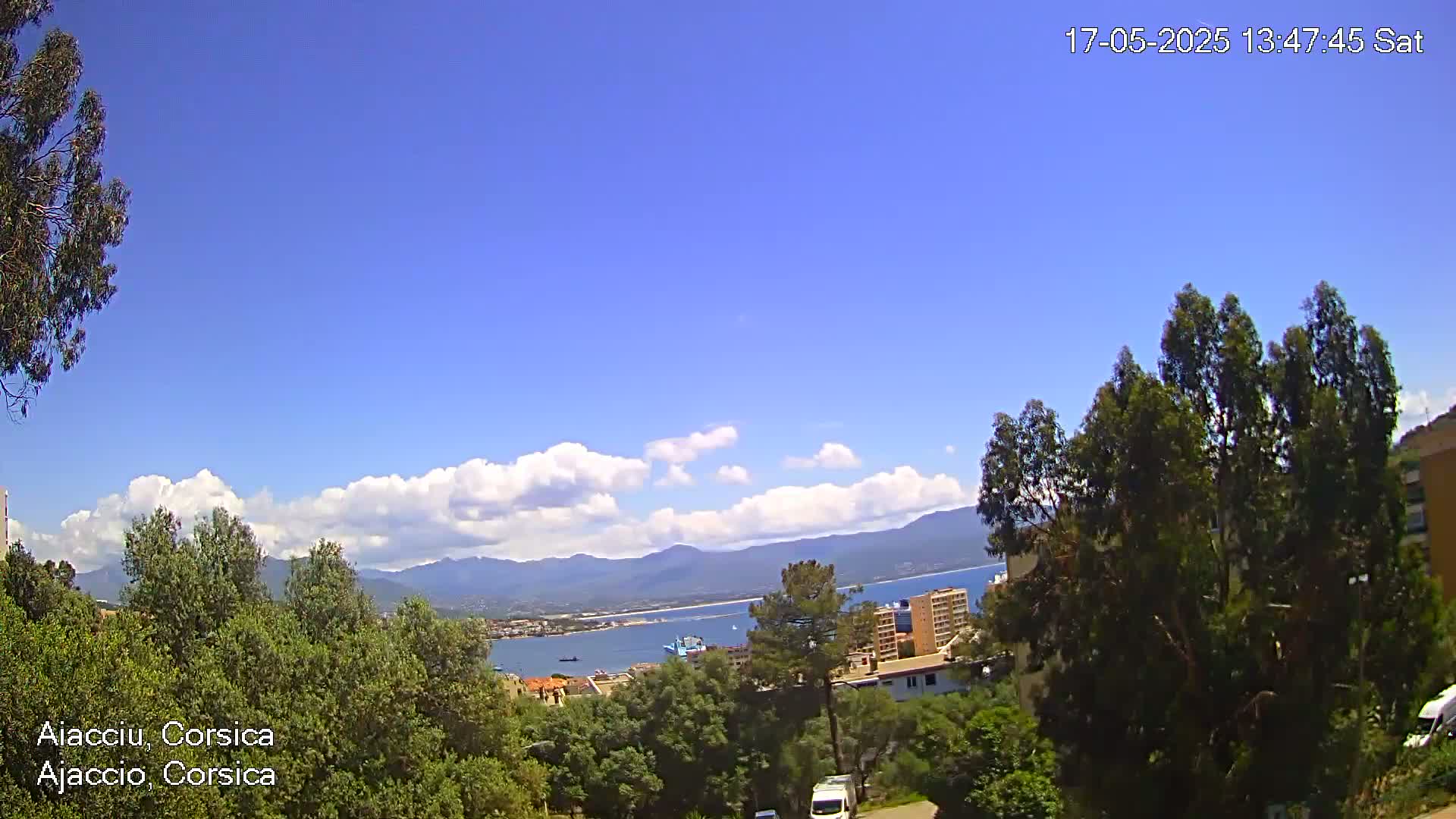 A partly sunny, clear day reveals a coastal city nestled among green hills and mountains, overlooking a calm bay with a few ships.
