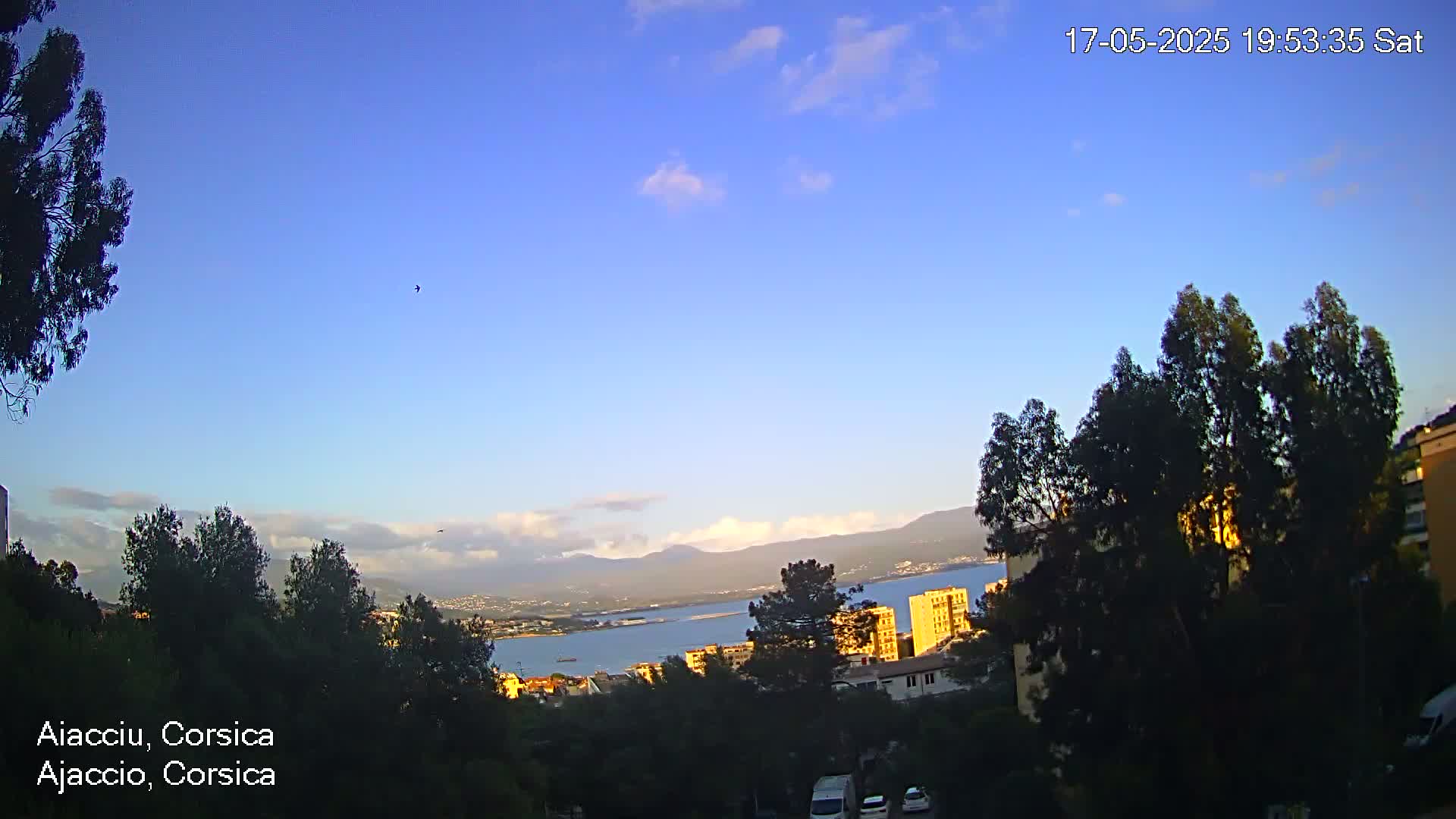 A partly cloudy, clear evening sky overlooks a coastal city with trees and buildings, and mountains visible in the distance.