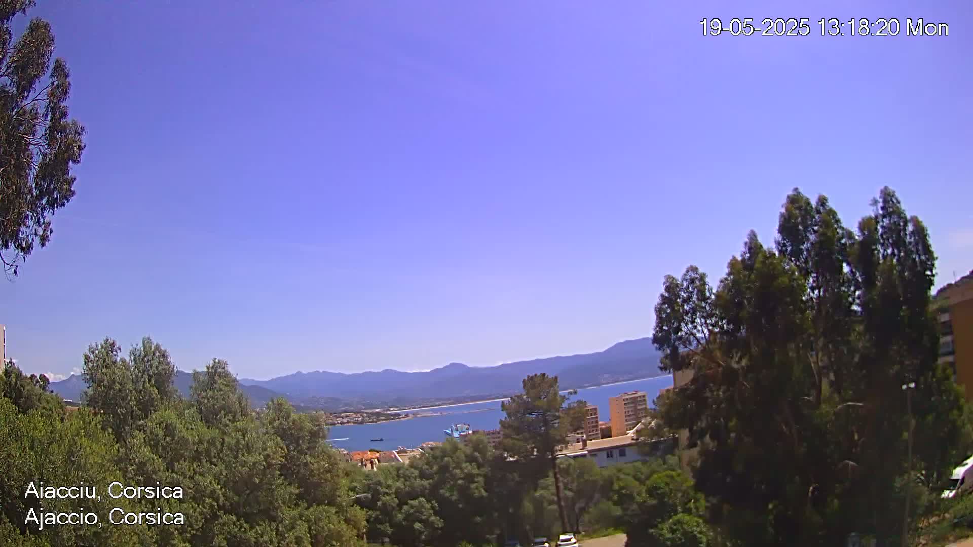 A clear, sunny day reveals a coastal scene with a calm body of water, mountains in the distance, and green vegetation in the foreground.