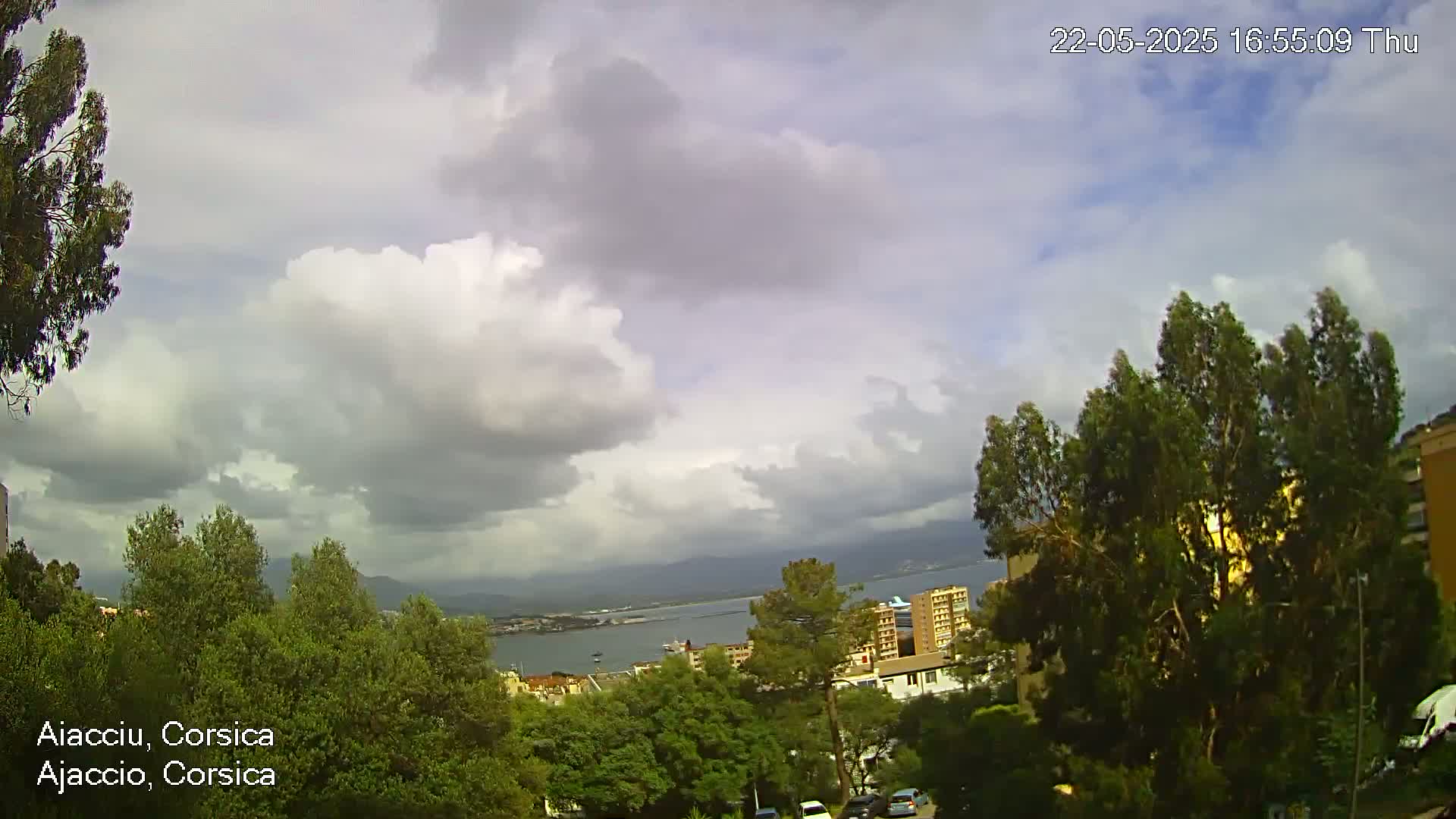 A partly cloudy sky overlooks a coastal city with green trees and a body of water in the distance.