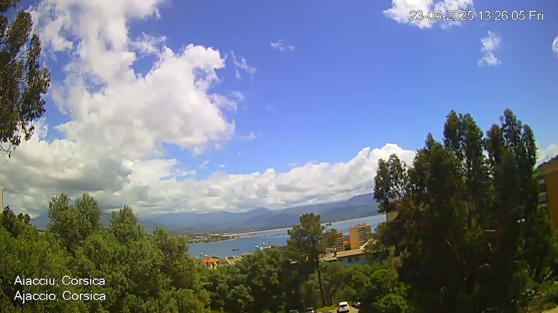 A partly cloudy day shows a view of a coastal town nestled among green trees and mountains, with a body of water visible in the distance.