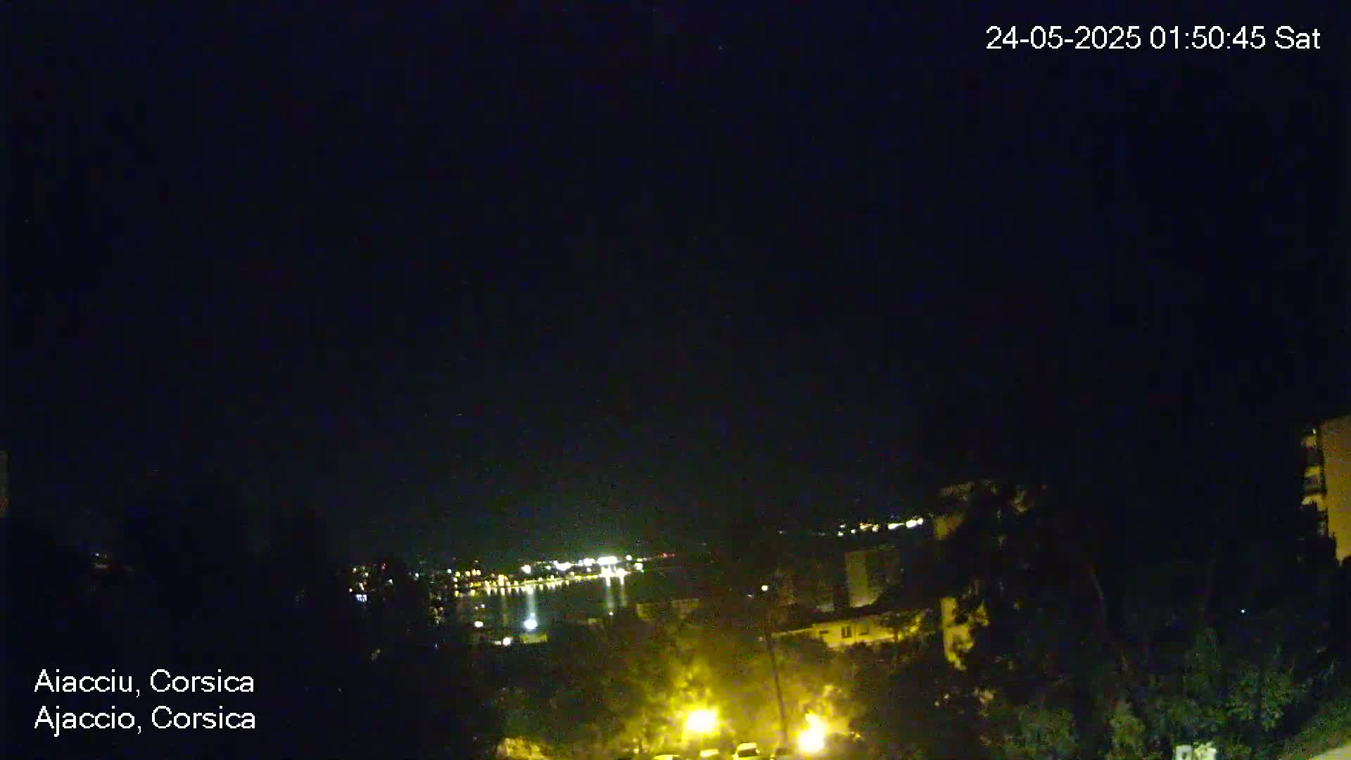 A nighttime view shows a dark body of water with distant lights, and a hillside with trees and illuminated streets in the foreground under a clear, dark sky.
