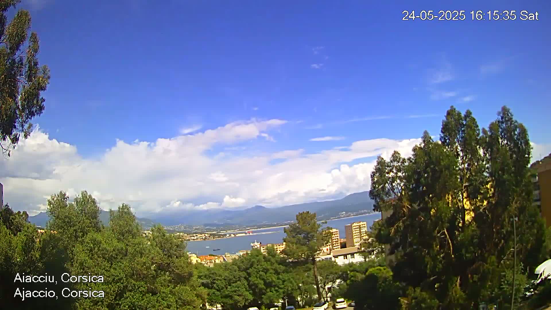 A partly cloudy blue sky overlooks a coastal town nestled amongst green trees and hills, with a body of water visible in the distance.